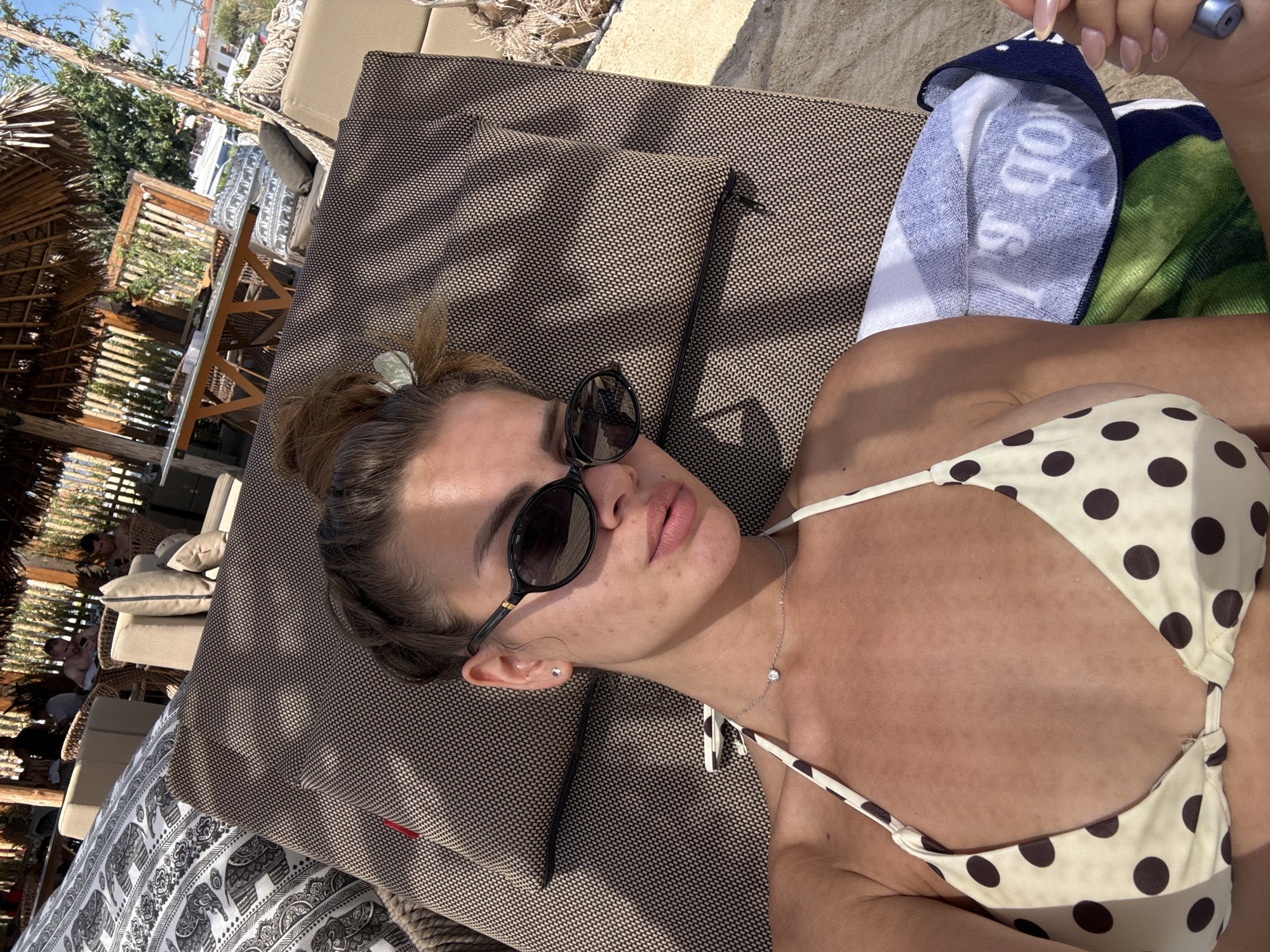 A young woman lies on a lounge chair at a beachside resort in Ofrynio Beach, Greece. She is wearing sunglasses and a polka dot bikini. Her hair is pulled up in a messy bun. A patterned cushion is behind her head, and a towel with blue and green stripes is draped over the chair. In the background, there are other lounge chairs, tables, and thatched roofs, indicating a resort or outdoor dining area. The sun is shining, casting shadows and highlighting the sand on the ground.
