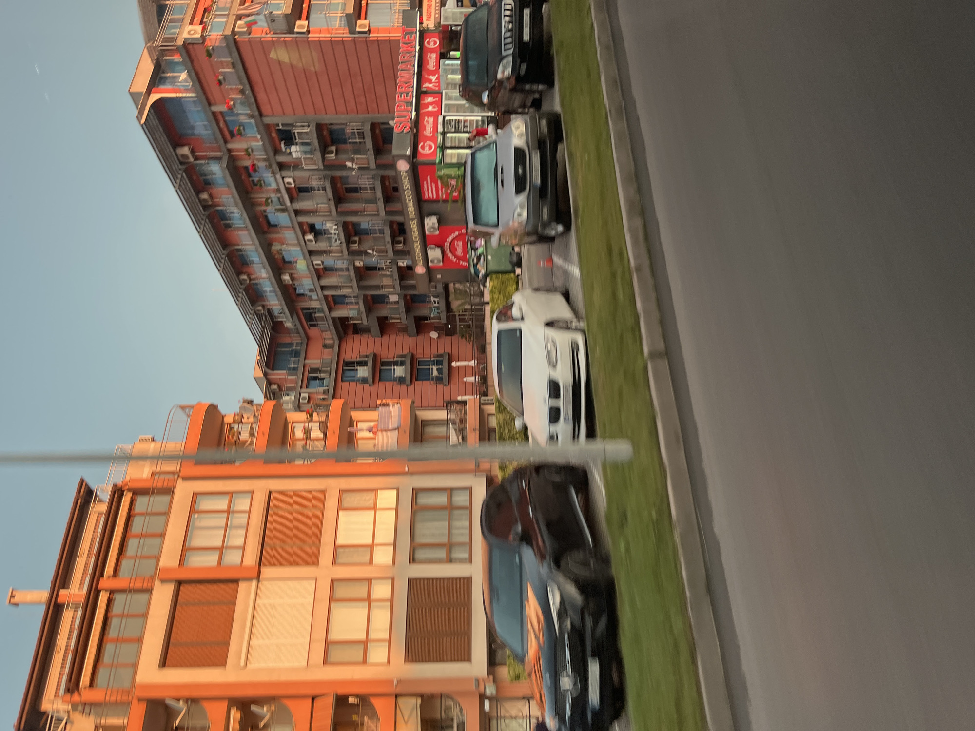 In Nesebar, Bulgaria, along a city street, a line of parked cars faces a multi-story building. The building, painted in shades of orange and cream, features numerous balconies and windows. To the right of this building, a lower structure with a red facade houses a "SUPERMARKET" and a "ALCOHOL CIGARS TOBACCO SHOP." Several vehicles, including a black Mercedes-Benz, a white BMW, a silver SUV, and a black SUV, are parked in front of the buildings. A streetlamp stands on the right side of the image. The sky is a clear blue, suggesting daytime.