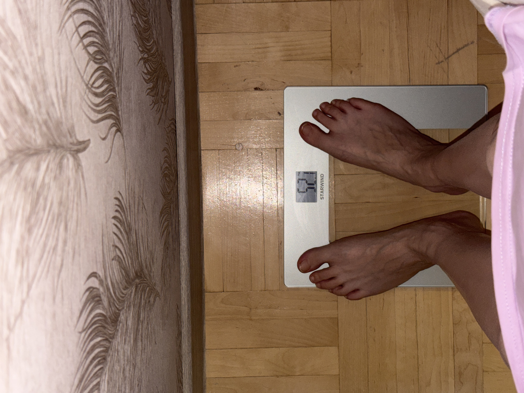 This overhead shot depicts a person's bare feet positioned on a white digital bathroom scale within an indoor setting in Izhevsk, Russia. The scale, bearing the brand name "STARWIND", clearly displays a weight reading of "62.1 kg".
The immediate surroundings indicate a home environment. The floor beneath the scale and feet is made of light-colored wooden planks, likely parquet or laminate. To the left, a wall covered in patterned wallpaper is visible, featuring a repeating design of what appears to be stylized feathers or leaves in muted tones. On the right side of the frame, a portion of a light pink garment, possibly pajamas or a dressing gown, hangs down, partially obscuring the lower right leg.
The lighting in the image is dim, suggesting an interior scene, possibly illuminated by artificial light rather than bright daylight. There are no direct cues about the time of day or outdoor weather. The focus is entirely on the act of weighing oneself, with no discernible emotions or other activities taking place. The composition emphasizes the scale and feet from a direct top-down perspective.