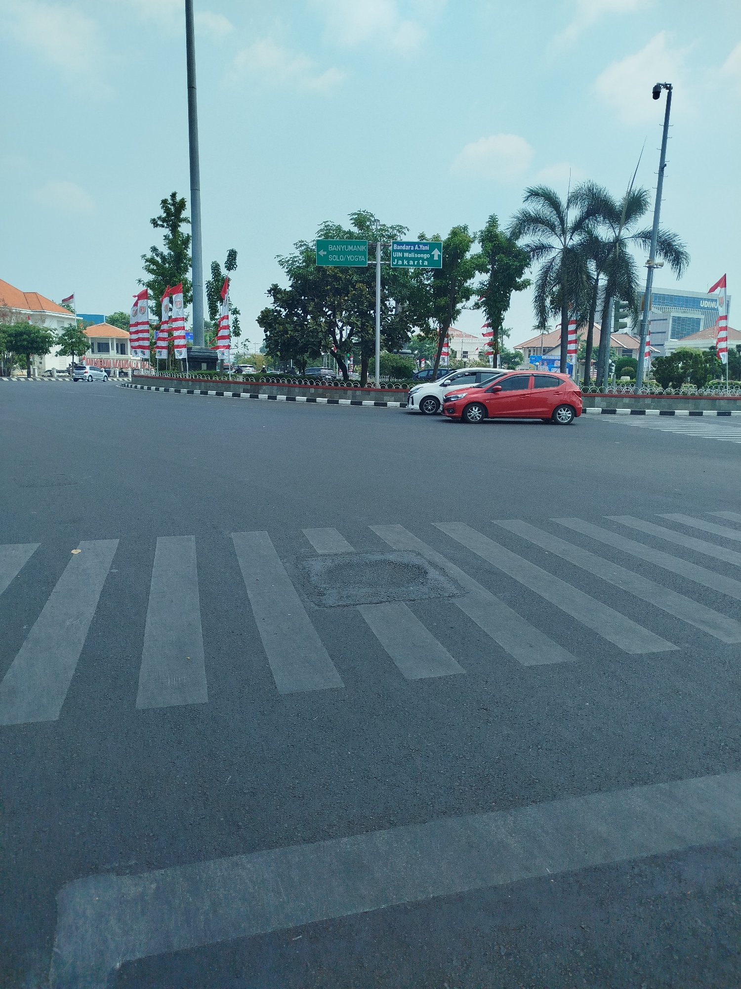 The image captures a street intersection in Semarang, Indonesia, under a bright, partly cloudy sky. A crosswalk with thick, white stripes dominates the foreground, marking a pedestrian crossing on the asphalt road. In the background, a white car and a red car are stopped at the intersection. To the left, a building with a red-tiled roof and white walls stands behind a row of decorative fences and several tall flags displaying red and white stripes. Beyond that, a large, grey flagpole is visible. Further right, palm trees and other foliage are interspersed with buildings, including a modern, multi-story structure with the word "UDINUS" visible on its facade. Green directional signs are mounted on poles; one points left with "BANYUMANIK SOLO/YOGYA" and another points up with "Bandara A. Yani UN Walisongo Jakarta." The overall scene suggests a busy urban environment, likely during daytime.