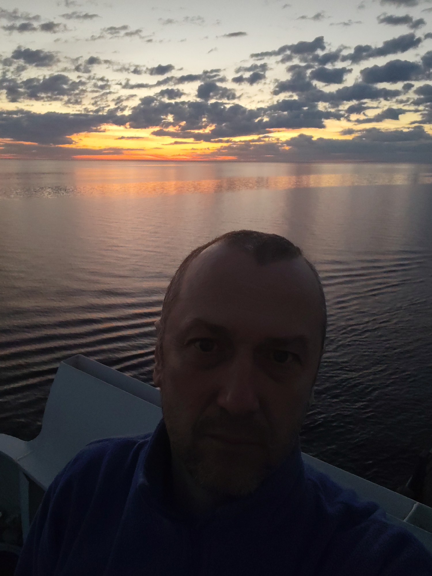 The photograph depicts a man taking a selfie against the backdrop of a sunset over the ocean. The man is in the foreground, wearing a blue top, and his face is clearly visible. Behind him is the ocean reflecting the orange hues of the setting sun. The sky has a mix of clouds, some dark and some illuminated by the sunset. There is a railing or structure of some sort visible, indicating he could be on a boat or a high vantage point. The location, Bol'shie Mkhi, Russia, is not directly evident from the visual elements of the image.