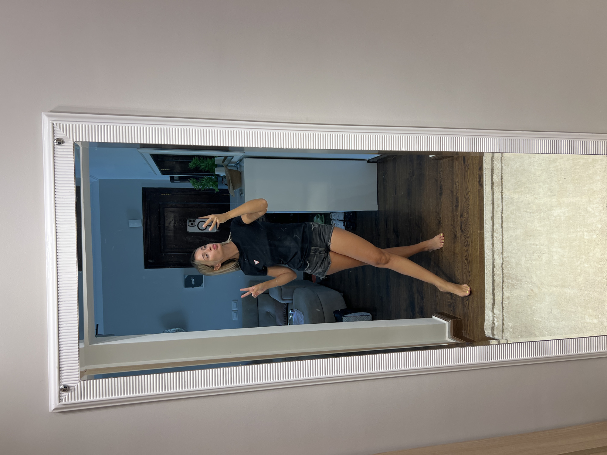 A woman is taking a selfie in a full-length mirror. She is wearing a black t-shirt and denim shorts. She is holding her phone in her right hand and making a peace sign with her left hand. Her lips are puckered. She is standing on a wooden floor in front of a beige rug. In the reflection, a living room area is visible with a couch and a dresser. The image appears to have been taken indoors, possibly in a residential setting. There is no visual information in the image that would allow for the determination of the city or country it was taken in, including Sharm El Sheikh, Egypt.