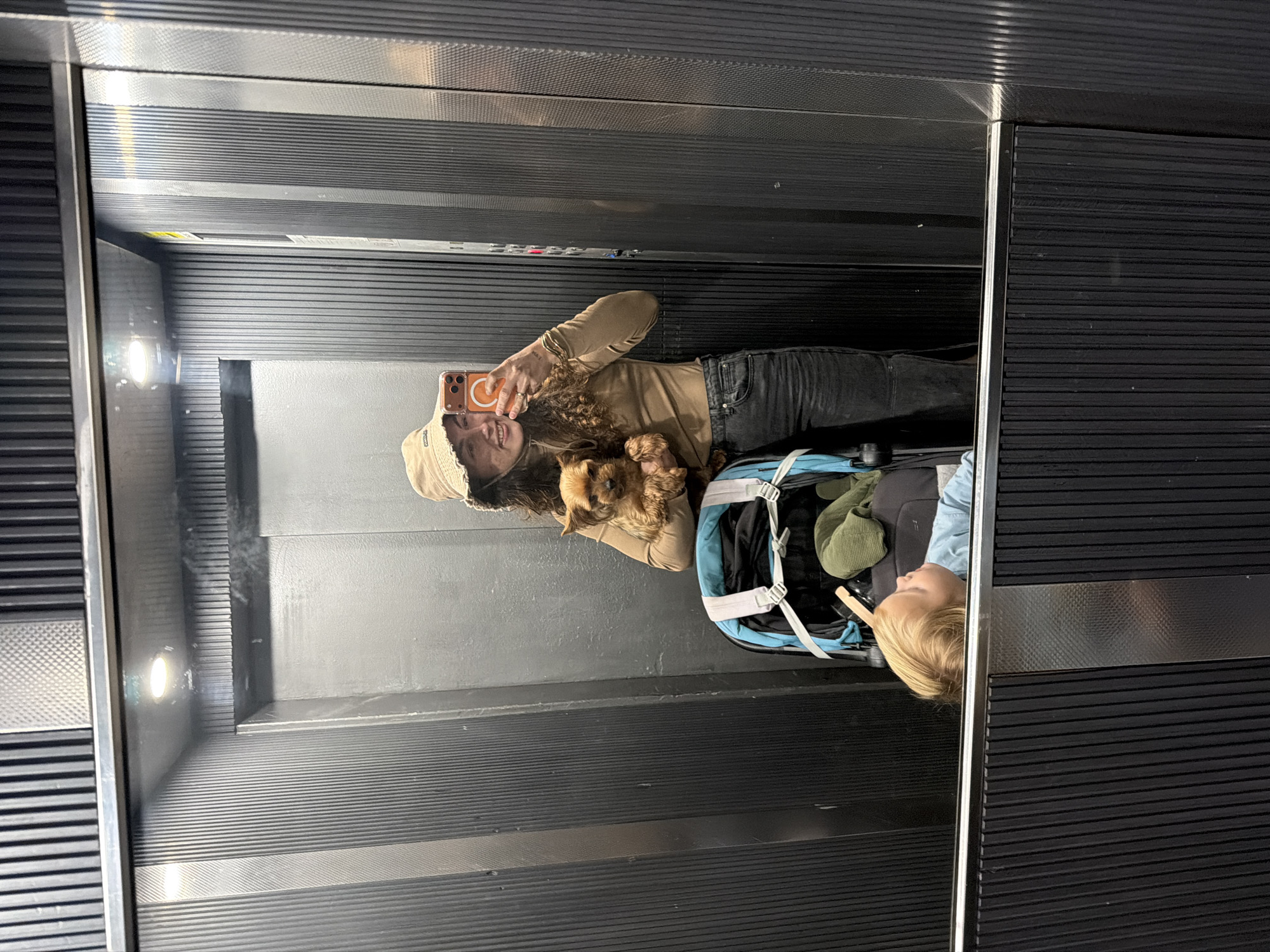 In an elevator with metal walls and ceiling, a woman in a tan bucket hat, brown long-sleeved shirt, and dark jeans is taking a selfie with her phone. She is holding a small brown dog. Beside her is a baby in a light blue shirt in a blue stroller. The elevator interior is characterized by horizontal grooves and metal paneling. The location is Kiryat Bialik, Israel.
