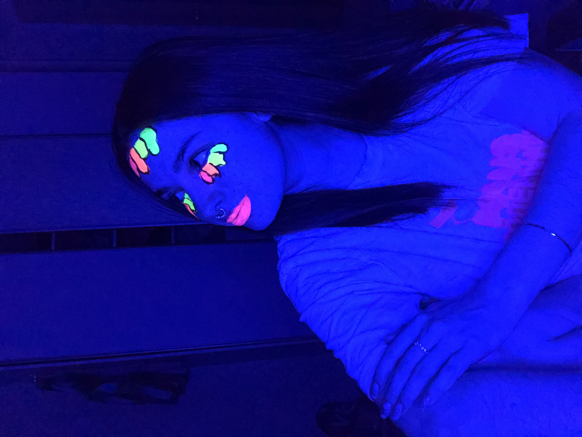 A young woman is seated with her hands clasped in her lap. Her face is adorned with fluorescent face paint, and she appears to be illuminated by ultraviolet lighting. The environment is dark, with a reflective surface visible below her. The location in the city of Noviy, Russia, is not determinable from the visual information provided.