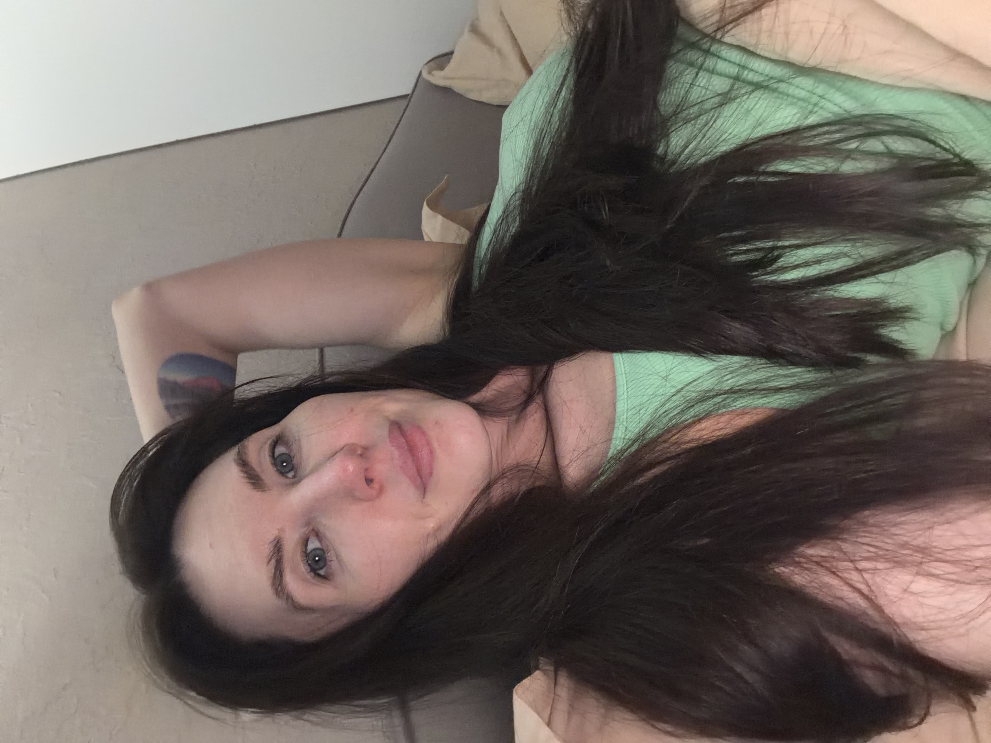 A woman with long dark hair and blue eyes is lying down, wearing a light green shirt. She has a tattoo on her upper arm. The location appears to be indoors, possibly on a couch. There is a wall and a beige object in the background. The city of Perm, Russia, is not discernible from the image.