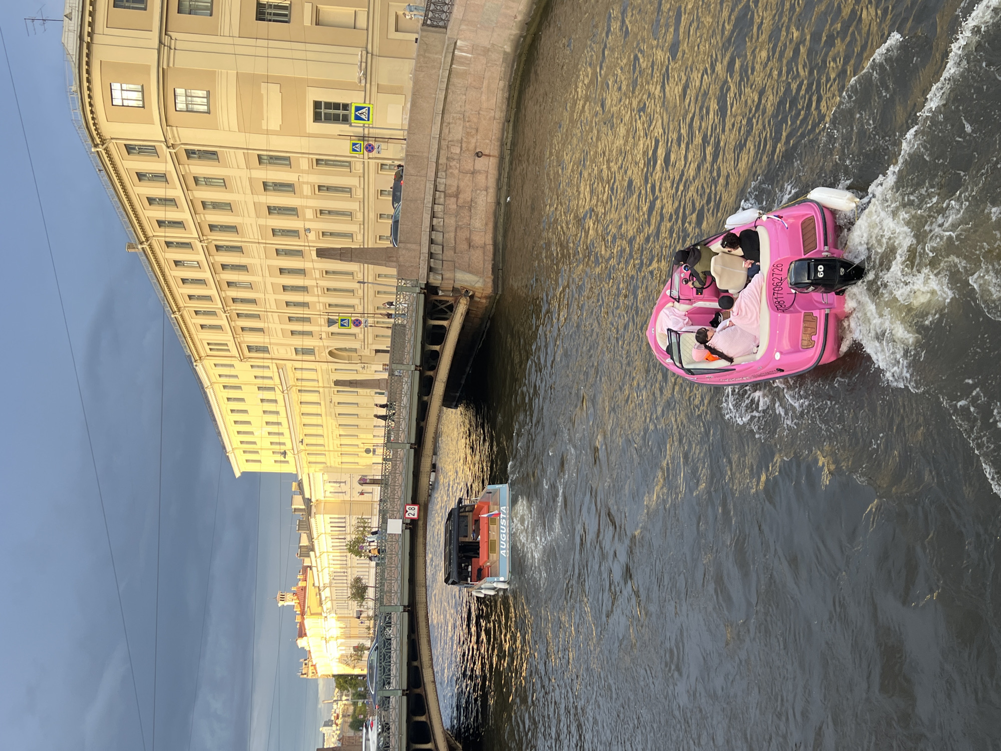 This media file captures a scene along a canal in Saint Petersburg, Russia. The dominant subjects are two boats on the water and the architecture lining the canal. The boat in the foreground is a vibrant pink, with several people on board. They appear to be on a tour or leisure outing. The second boat, further down the canal, is a lighter blue-gray color and is labeled "AUGUSTA." It also appears to be carrying passengers.
The setting is urban, with a large, light-colored building with many windows forming the backdrop on the left side of the image. The architecture suggests a historical European city. The canal water is murky and reflects the sunlight, creating a golden hue. The sky is partly cloudy, with a mix of blue and darker clouds, indicating either early morning or late afternoon. The presence of street signs with pedestrian crossing symbols ("A" inside a blue triangle) and what appears to be a street number sign ("28") on the bridge further confirm an urban setting.
The activity taking place is boat travel on the canal, likely a tourist activity given the nature of the boats and the presence of passengers. The pink boat is in motion, leaving a wake behind it. The people on board are casually dressed, and their expressions are not clearly discernible from this distance, but the overall mood appears relaxed.
Notable details include the bright pink color of the first boat, the name "AUGUSTA" on the second boat, the distinct architectural style of the buildings, and the play of light on the water. The time of day appears to be during daylight hours, with the sun casting long shadows and creating reflections. The weather seems to be fair, despite the presence of some clouds. The overall impression is a picturesque urban canal scene.
Visible text includes the number "60" on the outboard motor of the pink boat, the registration number "23817062726" on the side of the pink boat, and the word "AUGUSTA" on the side of the second boat. There are also traffic signs with the symbol of a pedestrian.