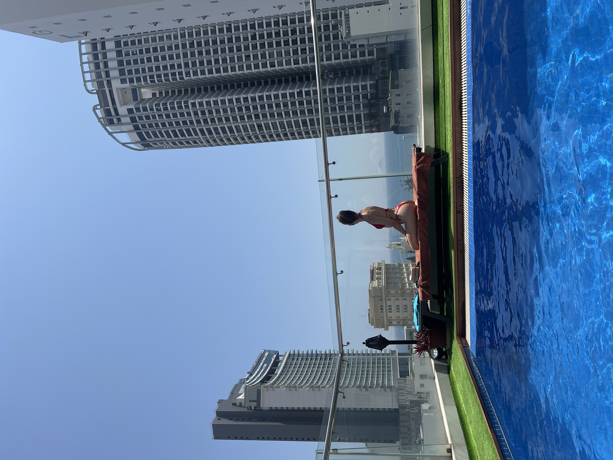 The image depicts a rooftop swimming pool area in Nha Trang, Vietnam, offering a panoramic view of the city and the South China Sea under a clear blue sky.
In the foreground, a bright blue swimming pool with rippling water occupies the lower half of the frame. The pool's edge is lined with light and dark blue tiles, wooden decking, and artificial green turf.
In the midground, a single female individual is seated on a sun lounger, seen from behind. She wears a red two-piece bikini, has her hair pulled back, and is facing away from the camera, looking towards the distant ocean and city skyline. The olive-green sun lounger is covered with a terracotta-colored towel. A light blue garment and a straw hat rest on the lounger beside her. A dark potted plant with reddish foliage and a black ornate lamp post are positioned near the lounger. A transparent glass railing with a metallic top rail separates the pool area from the drop-off.
The background reveals the cityscape of Nha Trang. On the left, a tall, modern building identified as the Hyatt Regency Nha Trang and Central Square features a distinctive curved facade of white, light grey, and dark grey sections. On the right, another prominent, multi-story building with a grid-like window pattern dominates the skyline. Beyond the buildings, the calm blue water of the South China Sea extends to the horizon, where a faint landmass and a small vessel are visible beneath a hazy sky. The sky above is cloudless and light blue.