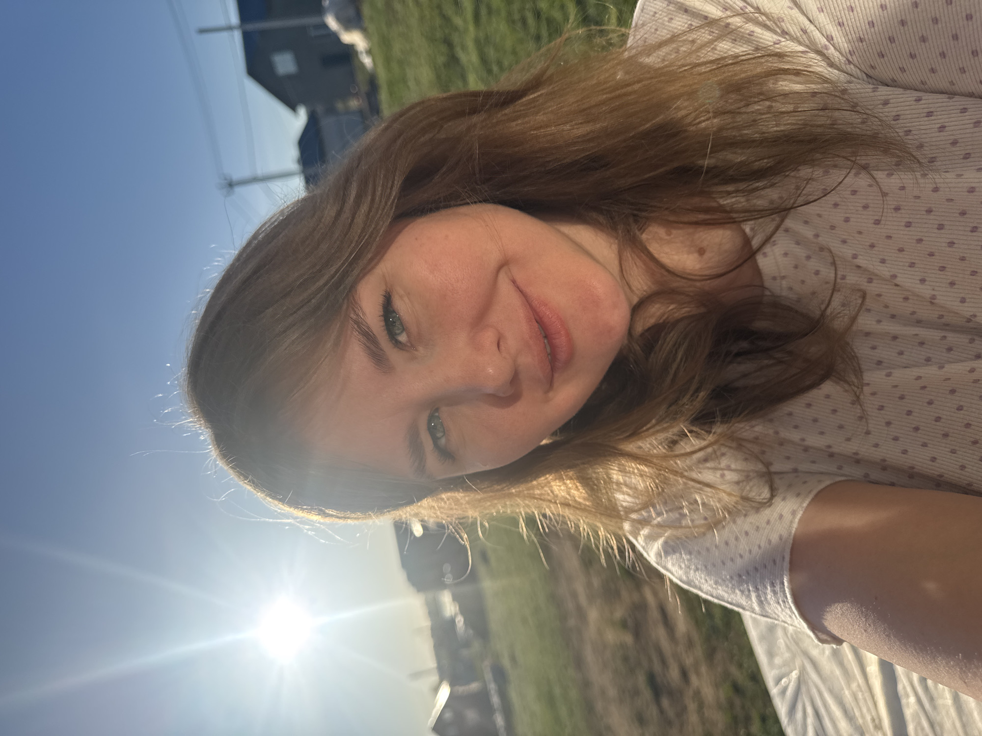 The image shows a selfie of a woman with fair skin and shoulder-length brown hair. She is smiling slightly and has light-colored eyes. She is wearing a light-colored shirt with small polka dots. The background includes a bright sun shining from the lower left of the frame, a grassy area, and a building in the distance. Based on the prompt, this scene is located in the city of Ферма, Russia.