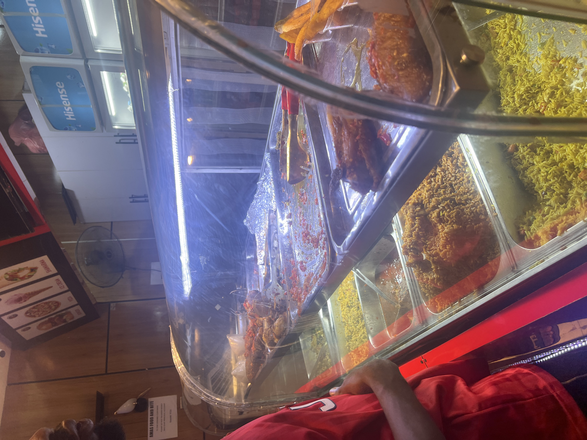 The image shows a partially visible man wearing a red shirt standing in front of a food display case. The display contains various dishes including rice, fried food, and what appears to be meat-based dishes. The man's arm and part of his head are visible in the lower part of the frame. The display case is brightly lit and reflects the environment. Behind the display, there are refrigerators labeled "Hisense," a wall fan, and a menu board listing items like Shawarma, Ice Cream, and Ribs. A sign reading "AMAS FOOD AND BITE" is also visible. Based on the sign, it's plausible that this scene is located in Abuja, Nigeria, where Amas Food is a known restaurant chain.