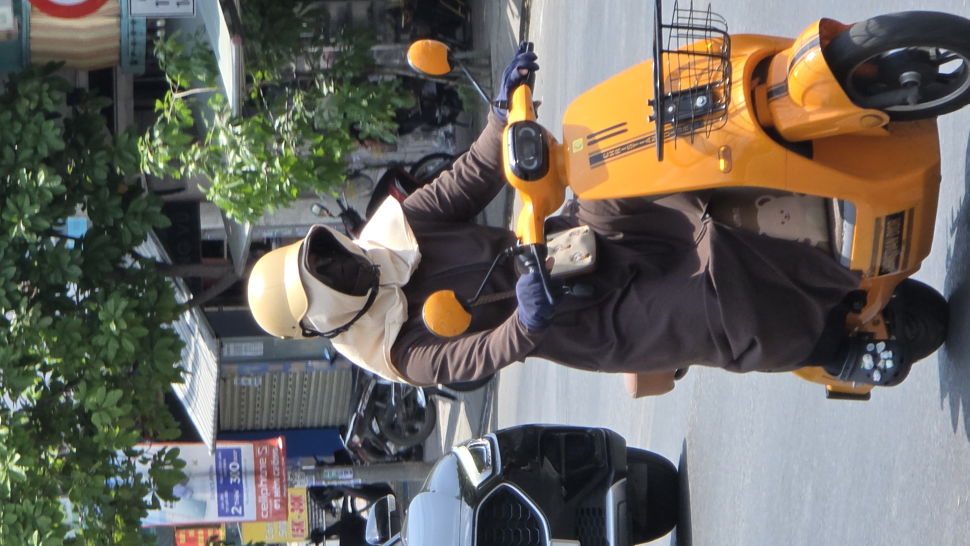 A person in a tan helmet and cream-colored head covering rides an orange electric scooter on a city street. The rider is wearing a brown long-sleeved shirt and blue gloves. The scooter has a black basket on the front and black stripes with white lettering on the side. In the background, there are parked scooters, storefronts, and trees. A black car is partially visible in the foreground. The image is taken during the day. It is located in the city of Bắc Nha Trang, Vietnam.