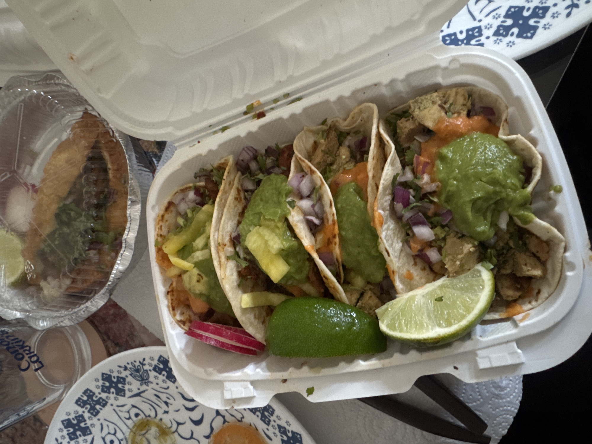 The image shows a styrofoam container filled with four tacos, topped with guacamole, red onions, and limes, along with a separate aluminum container that appears to hold additional food. The tacos appear to be a take-out order. There is also a dish with a blue and white pattern visible. This is a food scene; there is no indication of a specific location in New York City, USA.
