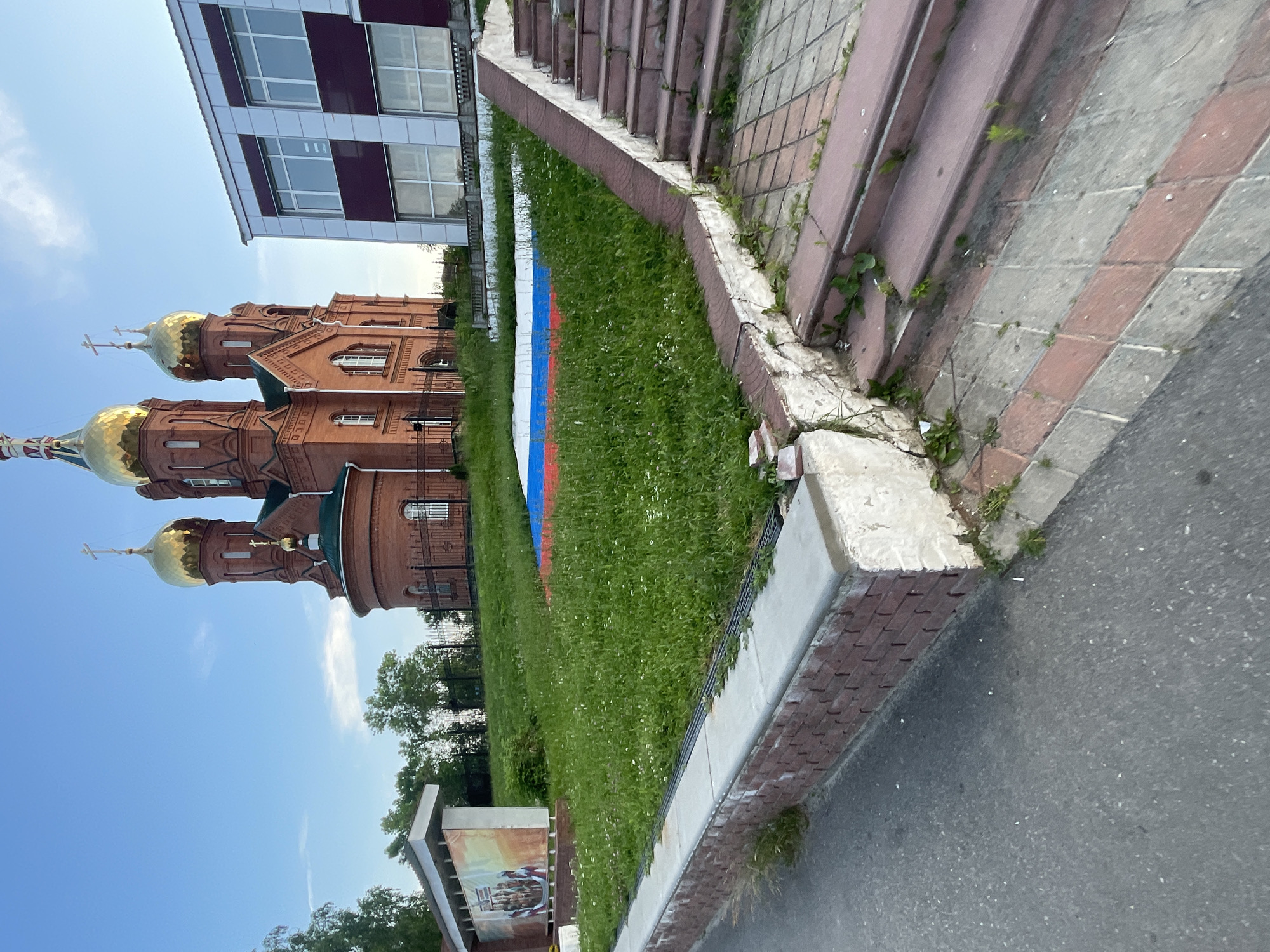 In the town of Krasnye Baki, Russia, a red brick church with golden domes stands prominently on a grassy hill under a clear blue sky. Adjacent to the church is a modern building with a facade of white and maroon panels. In front of the church, a large Russian flag is painted on the grass. A set of stone stairs with red brick accents leads up the hill towards the flag and the church. To the left of the church, a bus stop shelter with a mural depicting figures is visible. The scene is illuminated by natural daylight, suggesting it was captured during the day.