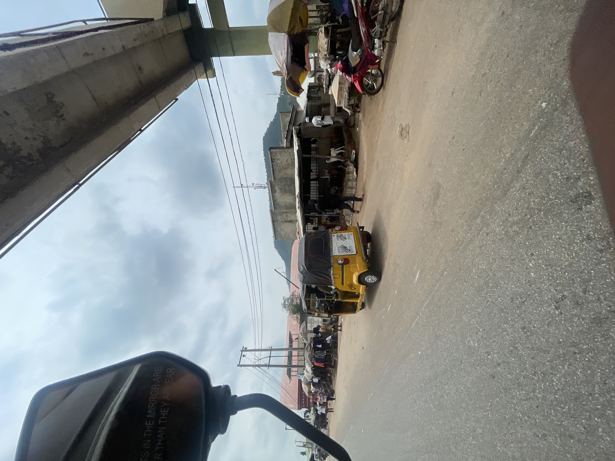 The image depicts a street scene in Lokoja, Nigeria, under an overcast sky. A yellow auto-rickshaw, also known as a Keke Napep, is visible in the middle of the road, with its passenger door open. To the right of the auto-rickshaw, a red motorcycle is parked near a market stall covered by an umbrella.
The market area features several stalls selling clothing displayed on racks, and some people are browsing. A group of goats can be seen in an enclosure behind one of the stalls. Further in the background, buildings and infrastructure, including what appears to be a section of an overpass or bridge, are present. Utility poles with numerous wires are also a prominent feature of the cityscape. The road surface is asphalt, and the overall impression is that of a busy, possibly informal, marketplace in an urban setting. The mirror of a vehicle is partially visible in the foreground on the left, showing text that reads "OBJECTS IN THE MIRROR ARE CLOSER THAN THEY APPEAR."
