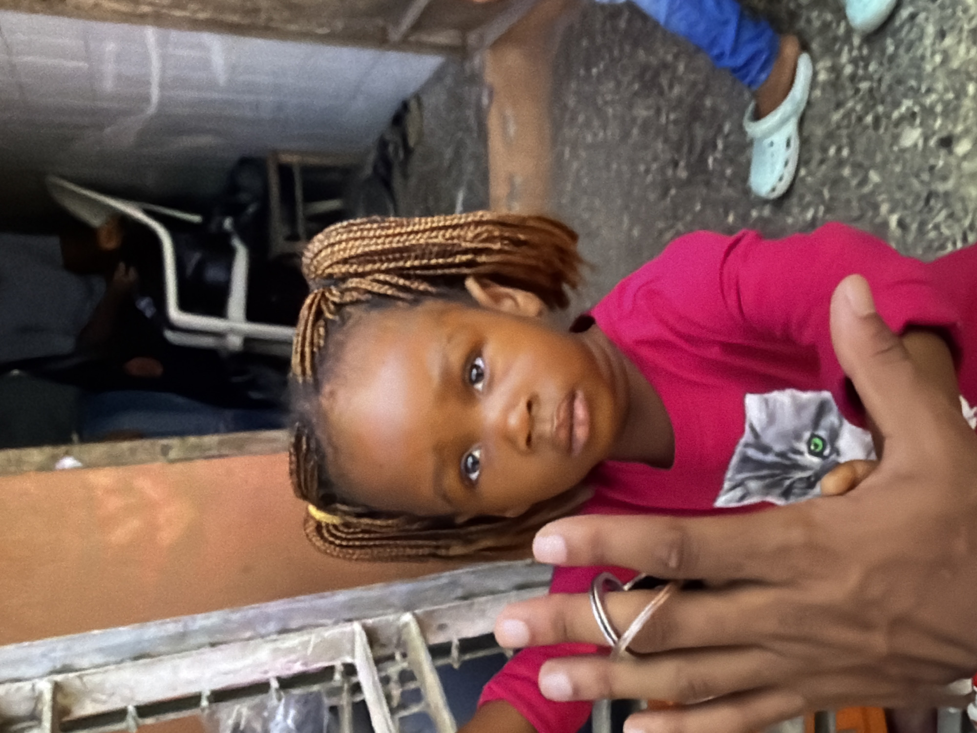 A medium shot captures a young Black girl with braided hair and a pink shirt featuring a cat graphic, looking directly at the camera. Her left hand, held up close to the lens, displays a silver ring on her ring finger. In the background, a person in blue pants and light-colored Crocs is visible, along with a white chair and a wall. The scene is set in an outdoor environment with a concrete floor and a textured wall. The lighting appears to be natural daylight, with some shadows indicating an overcast or partially shaded condition.