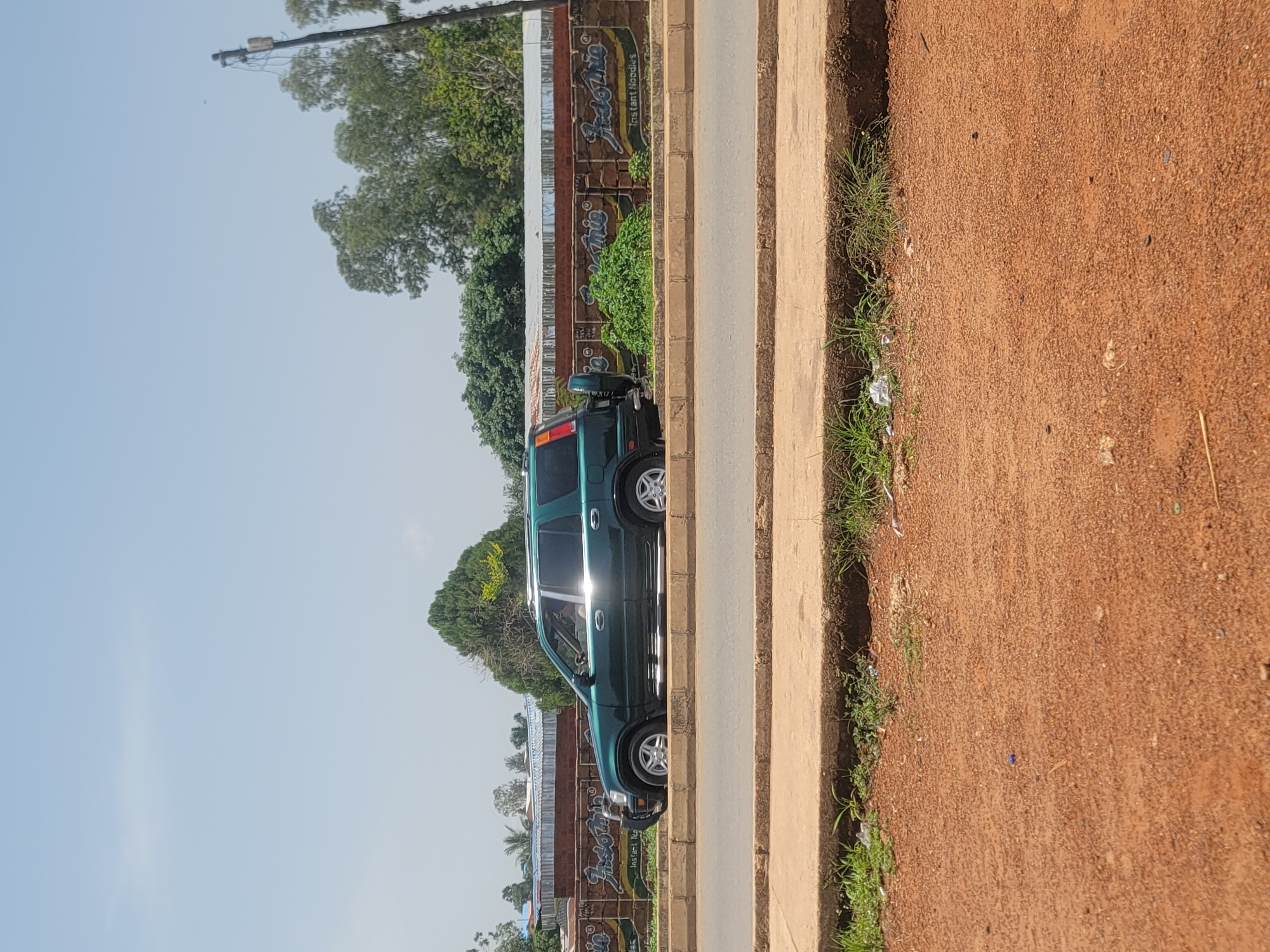 A dark green SUV is parked on the side of a paved road, parallel to a concrete curb. The SUV is a Honda CR-V with its spare tire mounted on the rear door. The vehicle is facing to the right of the frame and the driver's side is visible. Behind the SUV, a low brick wall runs along the edge of the road. This wall is decorated with graffiti that appears to advertise "Indo Mie Instant Noodles." Beyond the wall, there are trees and buildings, including what looks like a corrugated metal roof. The ground in the foreground is reddish-brown dirt with patches of dry grass and some scattered debris. The sky is clear and light blue. The scene suggests an outdoor setting in a town or city.