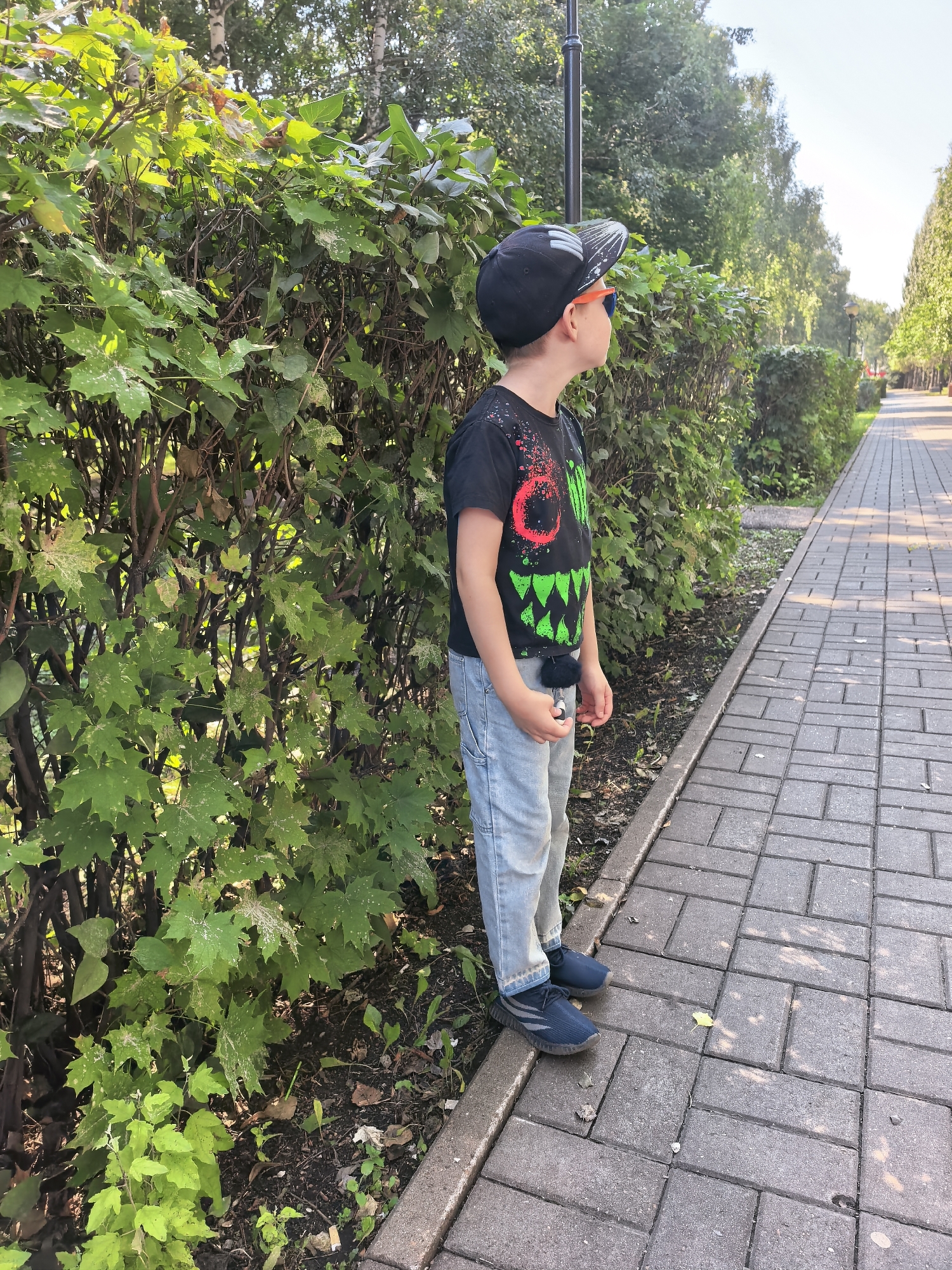 A young boy wearing a black t-shirt with a monster design, light blue jeans, a black baseball cap, and orange sunglasses stands on a paved sidewalk next to a dense green hedge. The boy is facing to the right, looking out of frame. A dark lamp post is visible behind him. The sidewalk is made of rectangular grey pavers and runs alongside the hedge, bordered by a curb. The background shows a tree-lined avenue with more hedges and trees. The scene appears to be a park or residential street in Sterlitamak, Russia, on a sunny day.