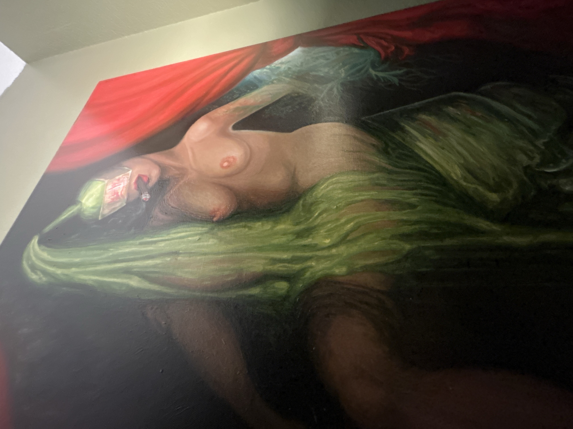 The image depicts an oil painting of a reclining nude female figure. She has pale skin and is draped in green fabric, with a red cloth partially covering her. She appears to be holding a smoking object near her face, possibly a cigarette. The background is dark, creating a dramatic contrast. The painting is hung on a wall, likely indoors, as indicated by the wall and ceiling visible at the top. There is no information available to locate the painting in Cape Coral, United States of America.