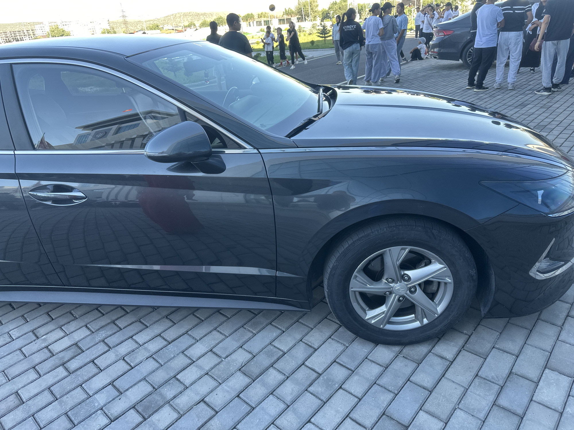 A dark gray sedan is parked on a paved area in front of a building. The car's front passenger side is visible, including the wheel, fender, door, and side mirror. The car appears to be a Hyundai Sonata, based on the wheel design and badge.
In the background, a group of people, including men, women, and children, are gathered. Some of the people are wearing casual clothing, and some appear to be playing with a basketball. The building behind them has a modern architectural style with multiple stories and large windows. The surrounding environment includes trees, a road, and a hilly landscape in the distance. The sky is clear and bright.
The image appears to have been taken in Manas, Kyrgyzstan, based on the inscription "KYRGYZ REPUBLIC" visible on the back of a black sweatshirt worn by one of the individuals in the background. The paved area suggests an outdoor public space, possibly a parking lot or plaza associated with the building. The lighting indicates it is daytime.