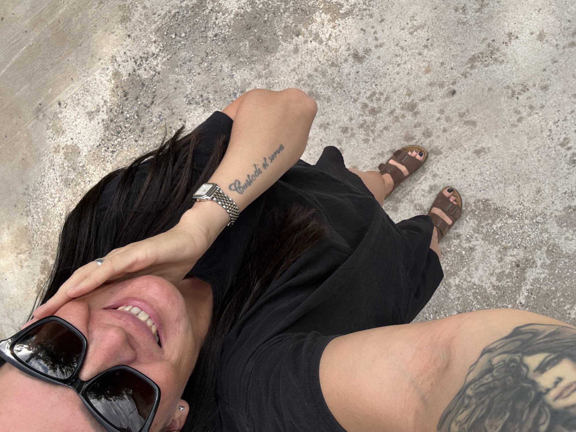 A woman is standing on a concrete surface. She is wearing a black t-shirt, brown sandals, and large black sunglasses. She has long dark hair and her right hand is on her face. There is a tattoo on her left forearm that reads "Custodi el serva" and another tattoo on her left upper arm of a woman's face. The concrete surface appears to be a sidewalk or pathway with small pebbles and debris. The image was taken from a high angle, looking down at the woman. The location is Chaikovsky, Russia.