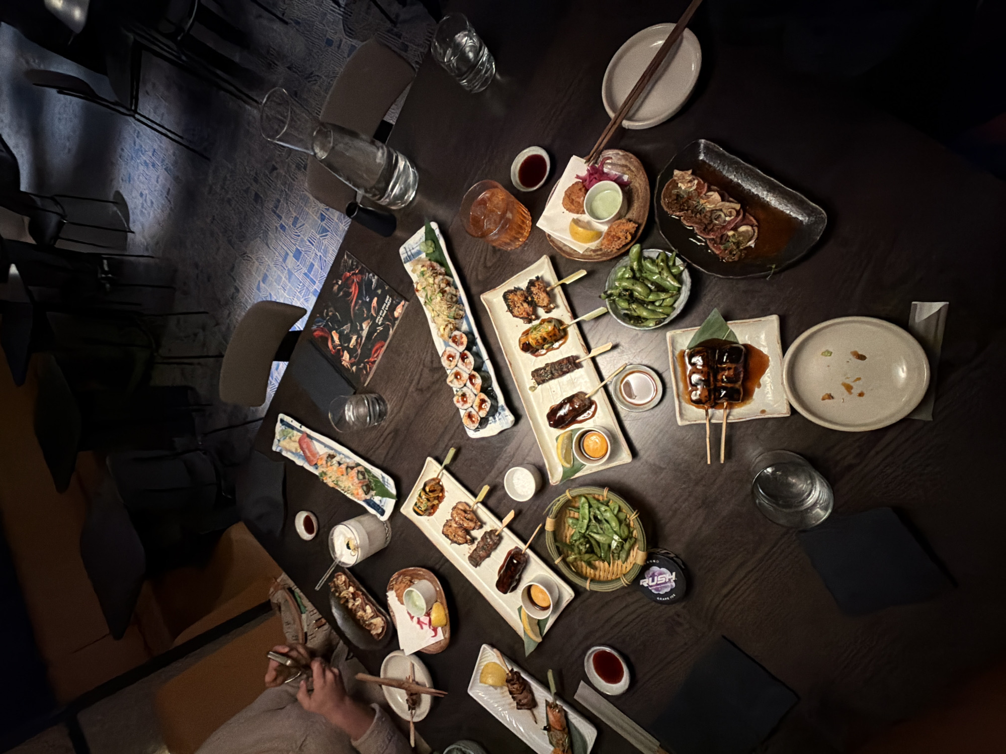 The image shows a table laden with Japanese cuisine, including sushi, skewers, and edamame, arranged on various plates and platters. Drinks, such as water and what appears to be a cocktail, are also present. Chopsticks are visible near some of the dishes. A person is seated at the table, holding chopsticks, possibly about to eat. The overall setting suggests a restaurant, with chairs visible around the table. The presence of a "RUSH" Grape Ice nicotine pouch indicates this photo was taken in London, United Kingdom.