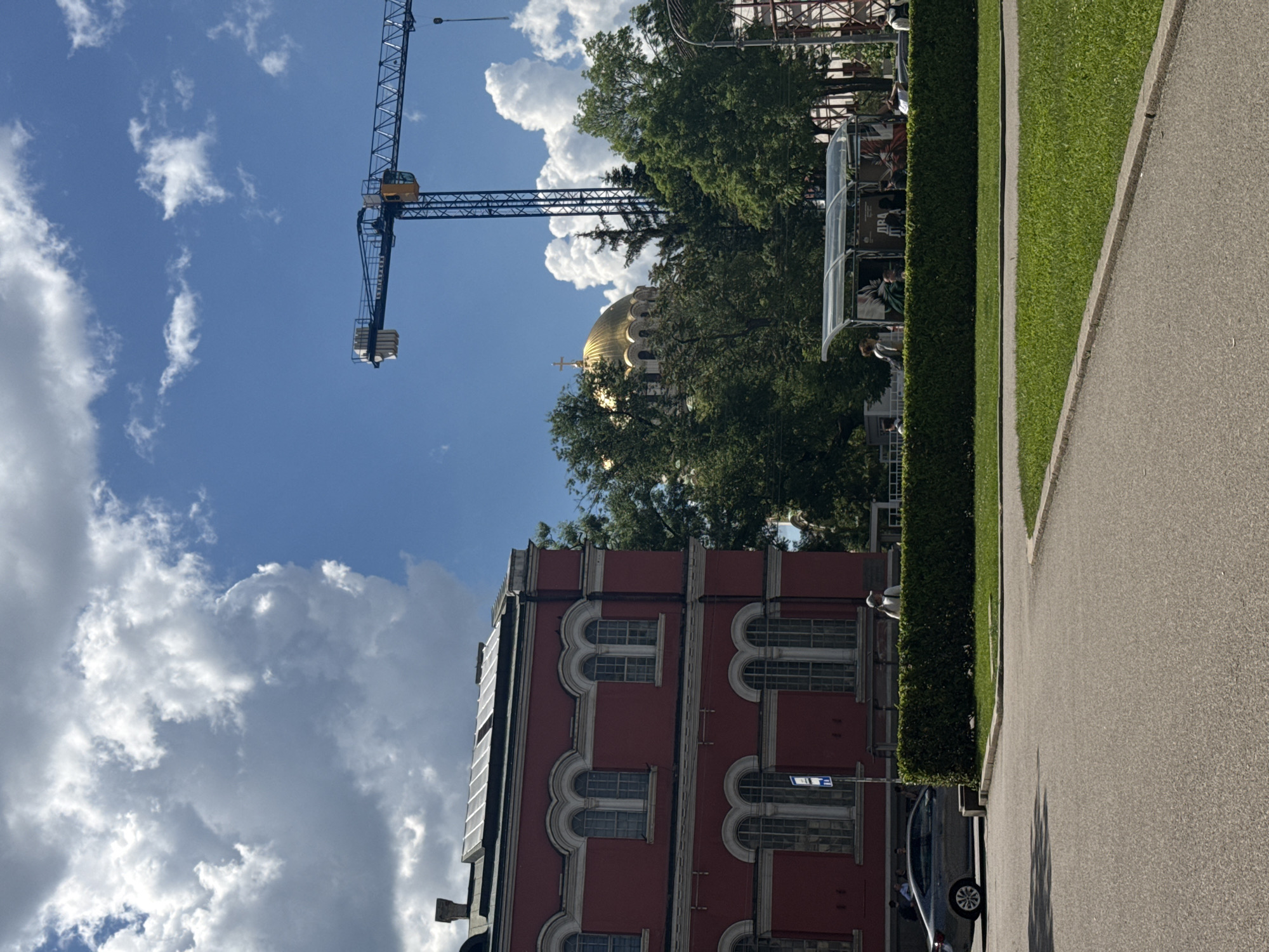 The image shows a street scene in Sofia, Bulgaria. A red building with multiple windows is in the lower-left portion of the frame. A golden dome is visible behind the building, partially obscured by trees. A construction crane is visible in the upper-left quadrant, standing against a blue sky with scattered clouds. A paved road, grass, and a trimmed hedge line the right side of the frame. There is a bus stop with people waiting inside. A car is parked along the road near the building.