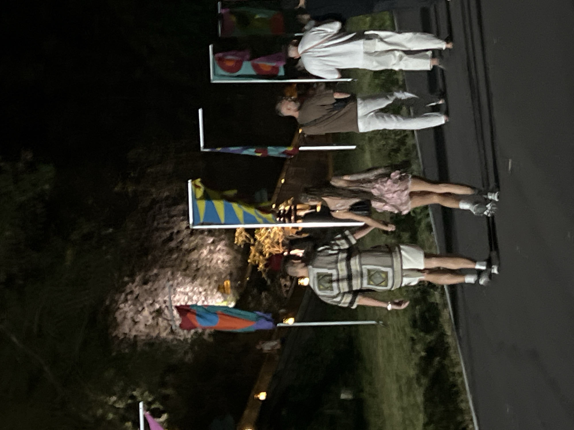 The image captures an outdoor gathering at night or dusk, with several people standing on or near a dark paved surface. In the foreground, people are dressed in casual attire; one individual wears a light pink dress and sneakers, while another sports a beige, brown, and black striped shirt with light shorts and sneakers, along with a watch. Further back, a bearded person in a brown shirt and light pants stands near another individual in a white shirt and white pants. The legs of additional people in white pants are also visible.
The scene is adorned with multiple colorful flags or banners on poles, featuring various geometric and abstract designs, such as blue and yellow diamonds, and combinations of red, orange, blue, and purple. Several light-colored rectangular structures, possibly display tables or vendor stands, are present, some holding red bowls or containers. The background consists of dimly lit trees and foliage, confirming an outdoor setting.
The activity appears to be a social event, likely a festival, fair, or outdoor market, where people are casually standing and mingling. The artificial lighting, though dim, illuminates parts of the scene, highlighting the individuals and flags against the dark evening backdrop. No specific emotions are clearly discernible from facial expressions, and there is no visible text in the image.