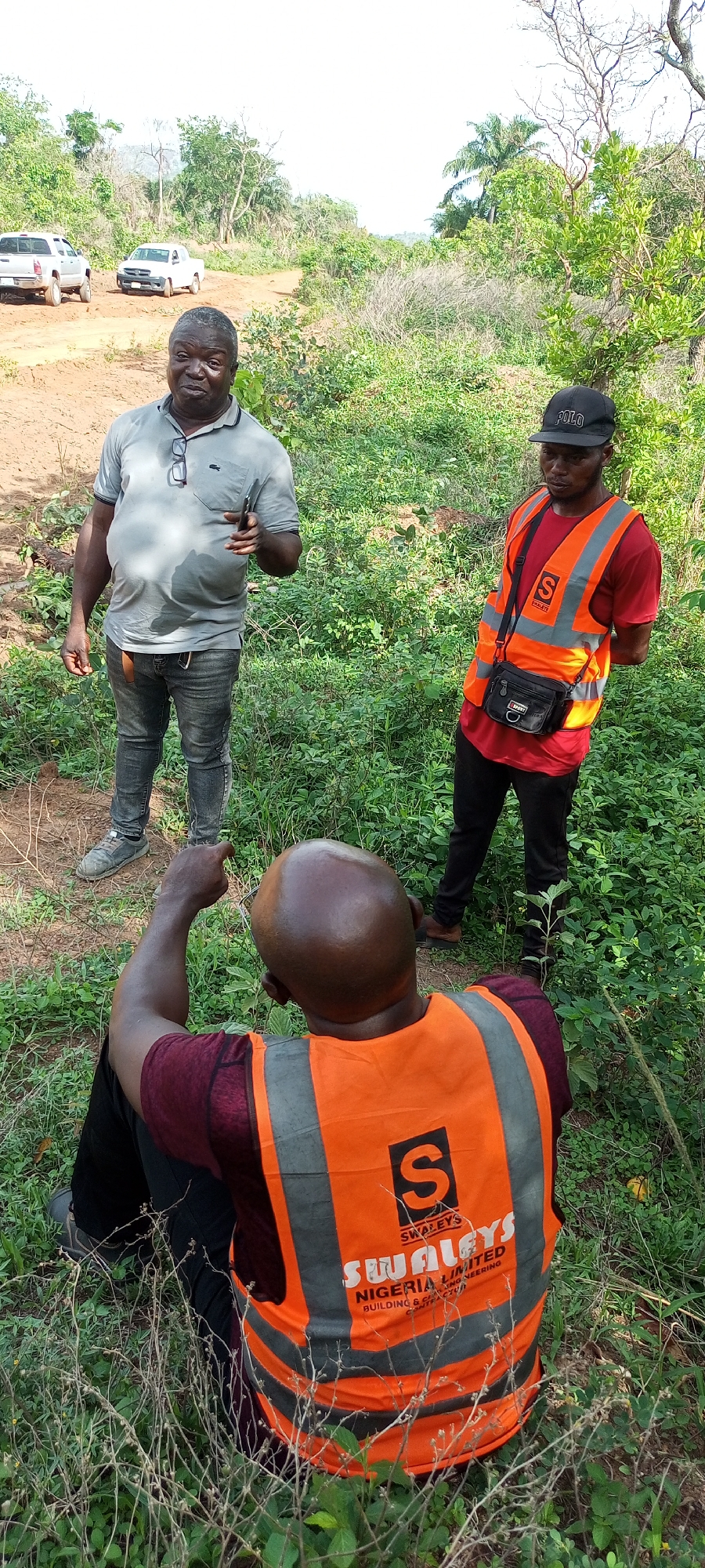 Three men are seen outdoors in a natural, possibly undeveloped, environment in Otunade, Nigeria, during daytime. The scene is dominated by lush green vegetation, including bushes, trees, and tall grasses, with a dirt track visible in the background where two white pickup trucks are parked.
In the foreground, a bald man with dark skin is seated on the ground, facing away from the camera. He wears a maroon long-sleeved shirt and an orange safety vest. On the back of his vest, a black logo with an 'S' and the text "SWALEYS NIGERIA LIMITED BUILDING & ENGINEERING CONTRACTOR" is visible. He is pointing forward with his right hand, seemingly directing attention to something.
Standing on the left, an older man with a sturdy build wears a light grey polo shirt, dark jeans, and grey sneakers. He has glasses hooked onto his shirt collar and holds a dark-colored phone in his right hand, gesturing as if speaking or explaining something.
To the right, another man stands, wearing a red t-shirt, black pants, and a black baseball cap with "POLO" written on it. He also wears an orange safety vest and a black shoulder bag across his body. He is looking down and slightly to his left, seemingly engaged in the discussion or observing what the seated man is pointing at.
The men appear to be involved in a professional discussion or survey of the area, possibly related to construction or development, given the safety vests and the visible company name on one of them. The lighting suggests it is bright daylight, possibly mid-morning or late afternoon.