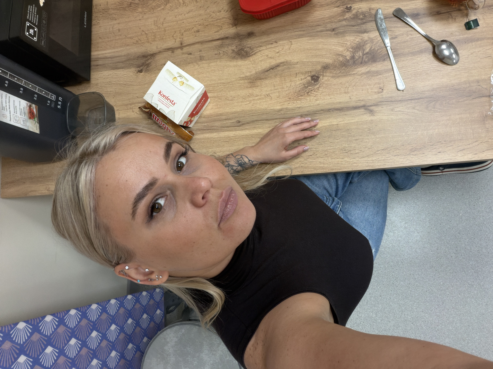 A woman is pictured in an overhead shot, positioned in front of a light wooden table. On the left of the table is a microwave oven and a water dispenser. In the upper left corner of the table there is a box of "Konfesta" chocolates and a "Denis" bar. A knife and spoon are on the right corner of the table. The woman has blonde hair and wears a black turtleneck sweater and blue jeans. Some of her skin is visible including her left arm which has a tattoo. A blue pattern from a chair appears in the lower left corner of the image and the gray floor of the room is visible. The woman is in Sarapul, Russia based on the text on the chocolate box.