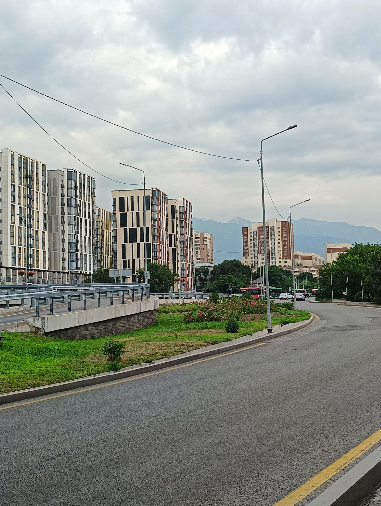 This media file depicts an urban scene in Almaty, Kazakhstan, under a cloudy sky. The foreground features a paved road with yellow lane markings curving to the right, bordered by a grass verge and a concrete barrier with a metal railing.
In the mid-ground, several modern, multi-story apartment buildings with patterned facades are visible. To the left, a raised section of road with a similar railing suggests an overpass or elevated roadway. Further down the road, traffic, including cars and a red bus, is moving. A green area with blooming pink flowers and small bushes separates the road from the buildings. Streetlights are spaced along the roadside.
In the background, more buildings of varying heights and architectural styles are present, interspersed with lush green trees. Beyond the city buildings, majestic mountains with snow-capped peaks can be seen against the overcast sky, indicating a relatively cool or temperate climate. The overall impression is that of a well-developed urban environment situated near a natural mountainous landscape. The lighting suggests it is daytime, but the heavy cloud cover implies overcast conditions, possibly before or after rain. There is no visible text in the image.