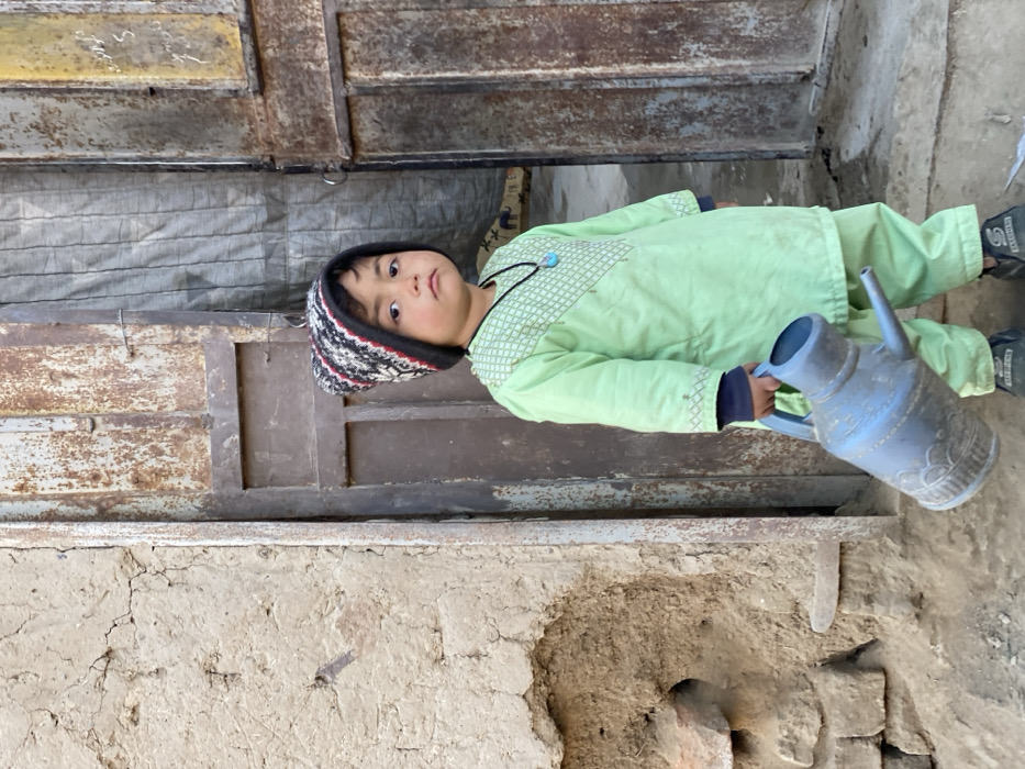 A young child stands in a dusty outdoor environment in Kandahar, Afghanistan. The child, wearing a multi-colored knit beanie, a light green long-sleeved tunic with embroidered geometric patterns on the chest and cuffs, matching light green trousers, and dark sneakers, holds a blue-grey plastic watering can with a molded decorative pattern in their right hand. A black cord necklace with a turquoise-colored pendant is visible on their chest. The child faces the viewer directly with a neutral expression. To the immediate left of the child is a weathered mud-brick wall exhibiting significant cracks and exposed brick material. To the child's right and extending behind them, two heavily rusted metal doors are set within a metal frame. The door on the right is ajar, revealing a darker interior space where a draped, textured grey fabric is visible. The ground beneath the child is dry, uneven dirt.