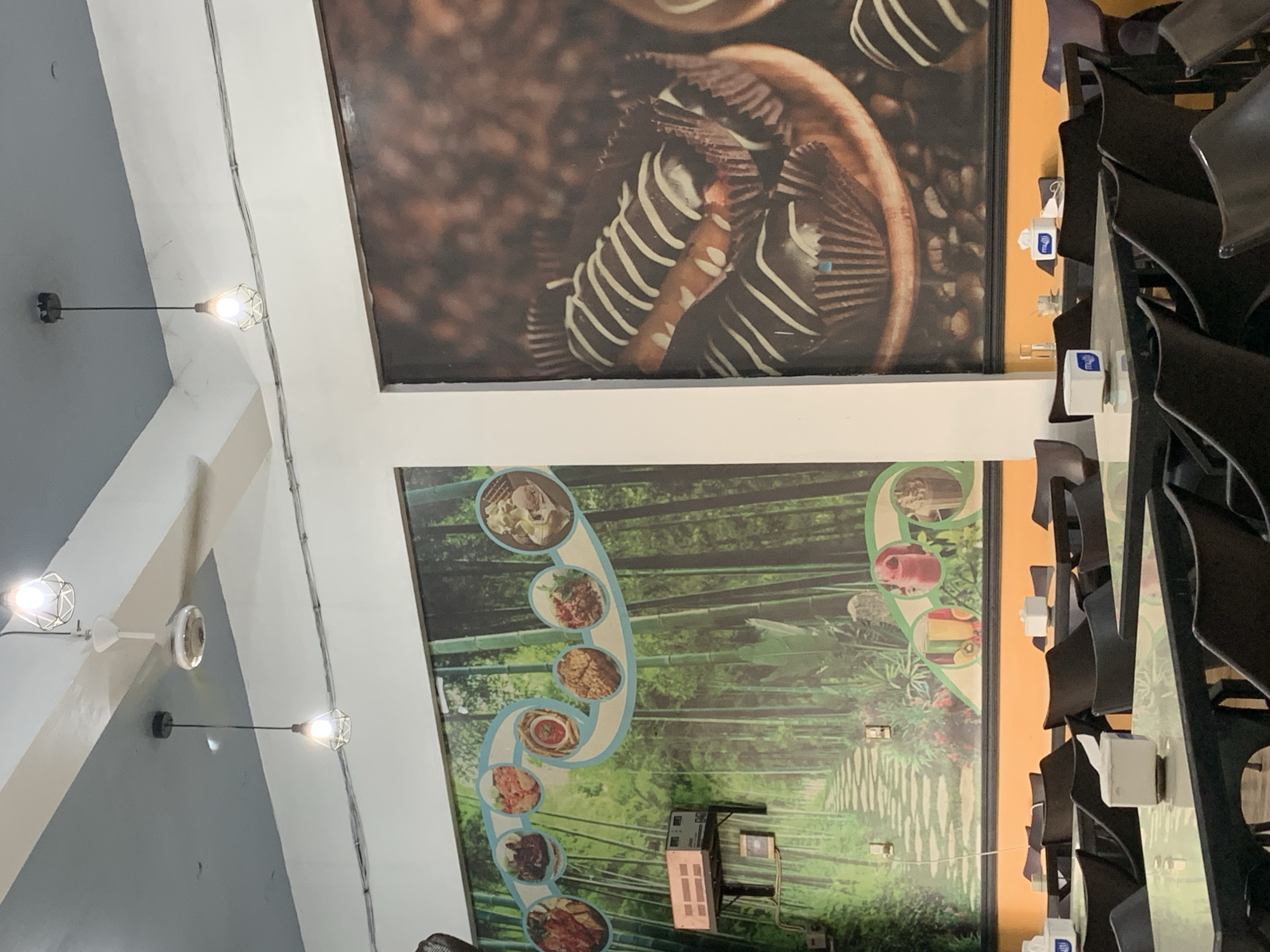 The image depicts the interior of a restaurant or cafe in Palohlada, Indonesia. The walls are decorated with large murals. On the left, a mural features a bamboo forest with a winding path and circular insets displaying various food items. On the right, a mural showcases a close-up of chocolate eclairs or similar pastries, set against a backdrop of coffee beans.
The restaurant appears to be set up with multiple tables and chairs. The tables are dark-colored with glass or reflective tops, and they are sparsely arranged with napkins and possibly tissue boxes. The chairs are dark brown or black and are of a simple, modern design.
Overhead, there are lighting fixtures. One is a white ceiling fan, and two others are pendant lights with wire cage-like shades, emitting a warm glow. The ceiling is painted in two tones, gray and white, and there are electrical conduits running along the wall.
The overall ambiance suggests a casual dining establishment. There are no people visible in the image, so no actions or interactions are depicted. The environment is indoors, with artificial lighting.