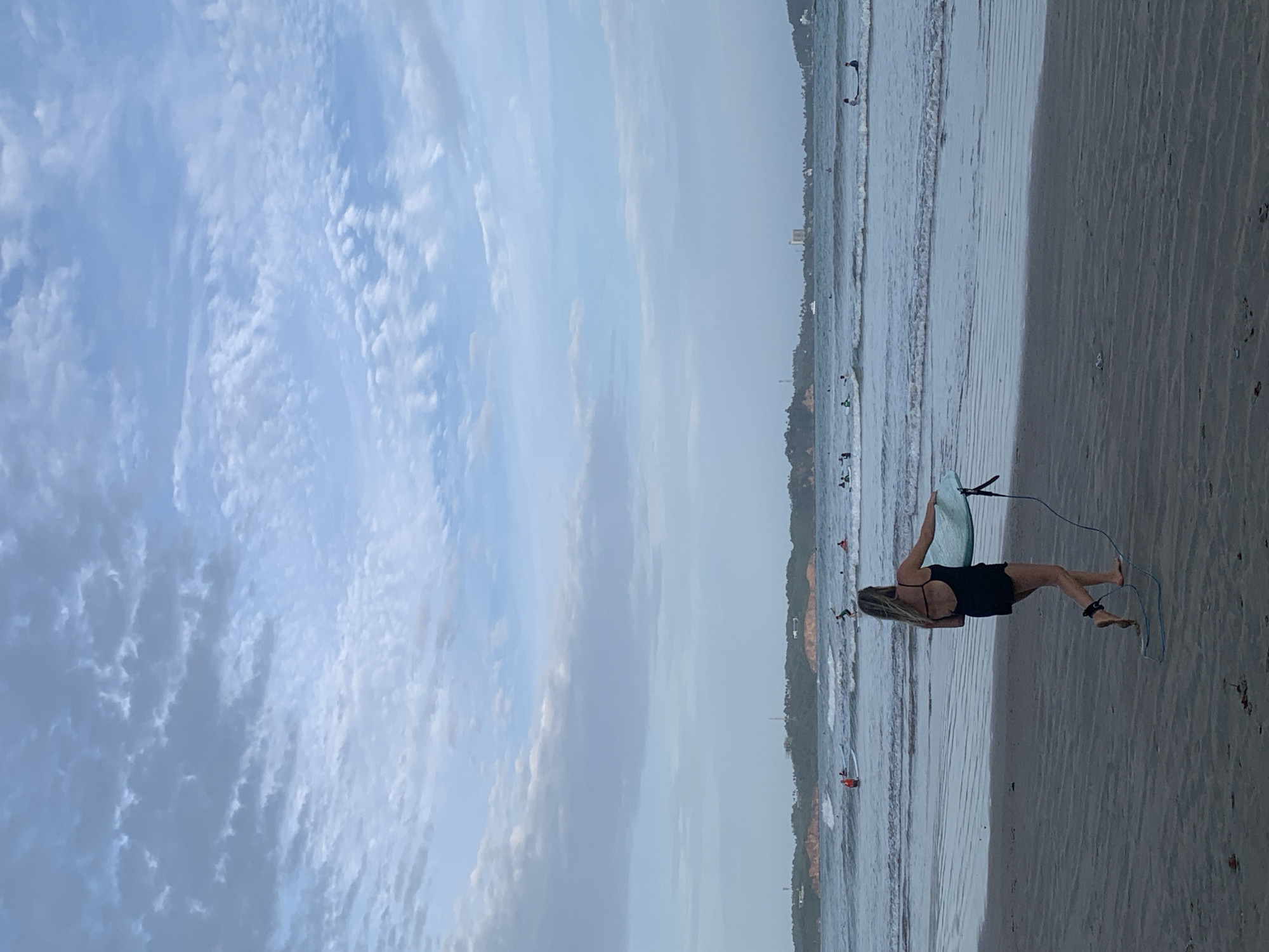 A full outdoor shot shows a woman standing on a beach with a surfboard, presumably in Weligama, Sri Lanka. The woman has long blonde hair and is wearing a black swimsuit. She is holding a light blue surfboard with black straps. The board is vertical, positioned between the water and her right side. The shoreline has waves. There are other people and boards visible in the water, some possibly windsurfing. The background shows coastal vegetation and possibly buildings. The sky has a variety of white and grey clouds.