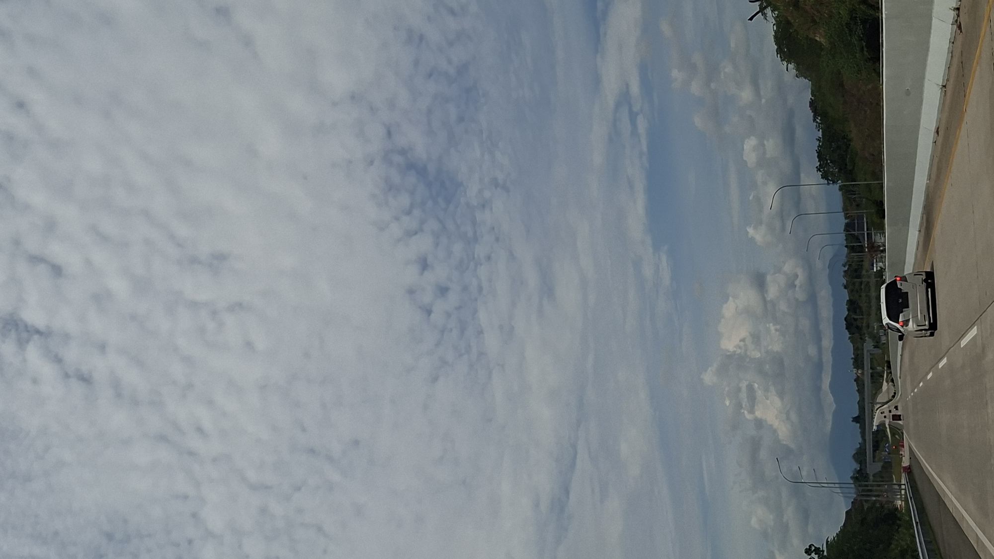 The image shows a view from a car driving on a highway in Keureuweungkrueng, Indonesia. The sky is filled with fluffy white clouds. The highway stretches ahead, with a white car in the right lane. Trees and hills are visible on the sides of the road, with mountains in the distance. The overall impression is of a clear, sunny day on a scenic drive.