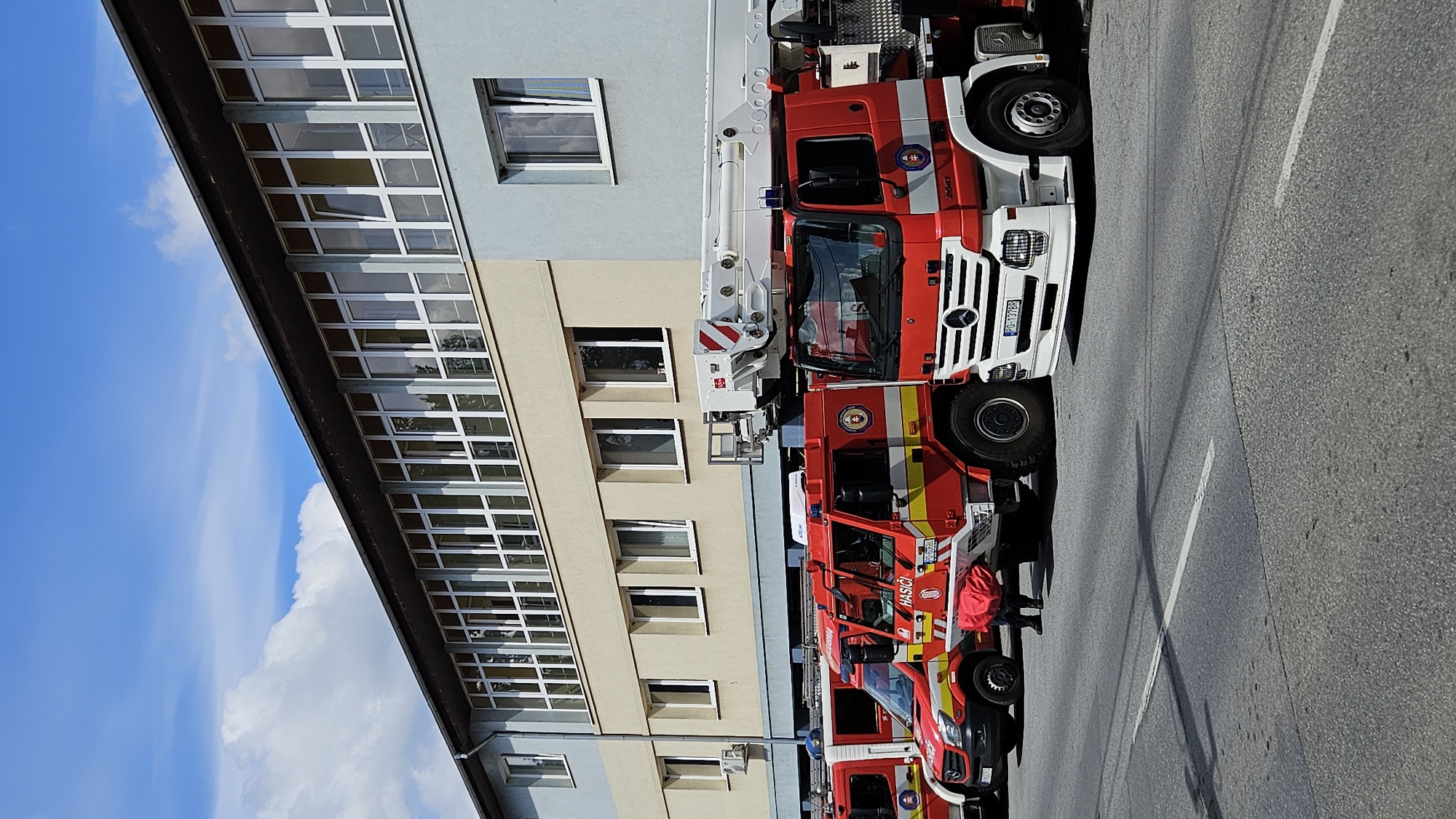 The image depicts a row of fire trucks parked on a paved road in front of a light-colored building with a blue and gray lower facade. The building has a long row of windows across its upper section and several individual windows on the lower sections. The sky is blue with scattered white clouds.
The fire trucks are primarily red and yellow, with some white accents. The truck on the far right is a Mercedes-Benz truck with a license plate reading "PO-137BR". The truck next to it has a white extendable ladder mounted on top. Another fire truck visible has "HASICI" written on its side. One person, dressed in dark clothing, is partially visible near the rear of one of the fire trucks, appearing to interact with a red object.
The context of this image is a fire station in the city of Prešov, Slovakia. This is inferred from the presence of multiple fire trucks, the "HASICI" marking (which translates to "firefighters" in Slovak), and the license plate format which is consistent with Slovakian plates.