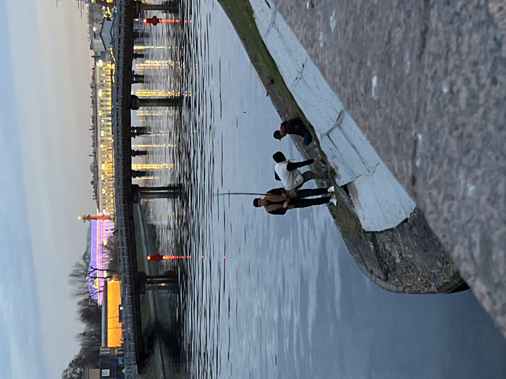 Three people are fishing from a concrete embankment. A man is holding a fishing rod. Buildings are in the background. A bridge with lights is visible over the water. The sky is dusky. This appears to be Saint Petersburg, Russia, based on the architectural styles and waterways.