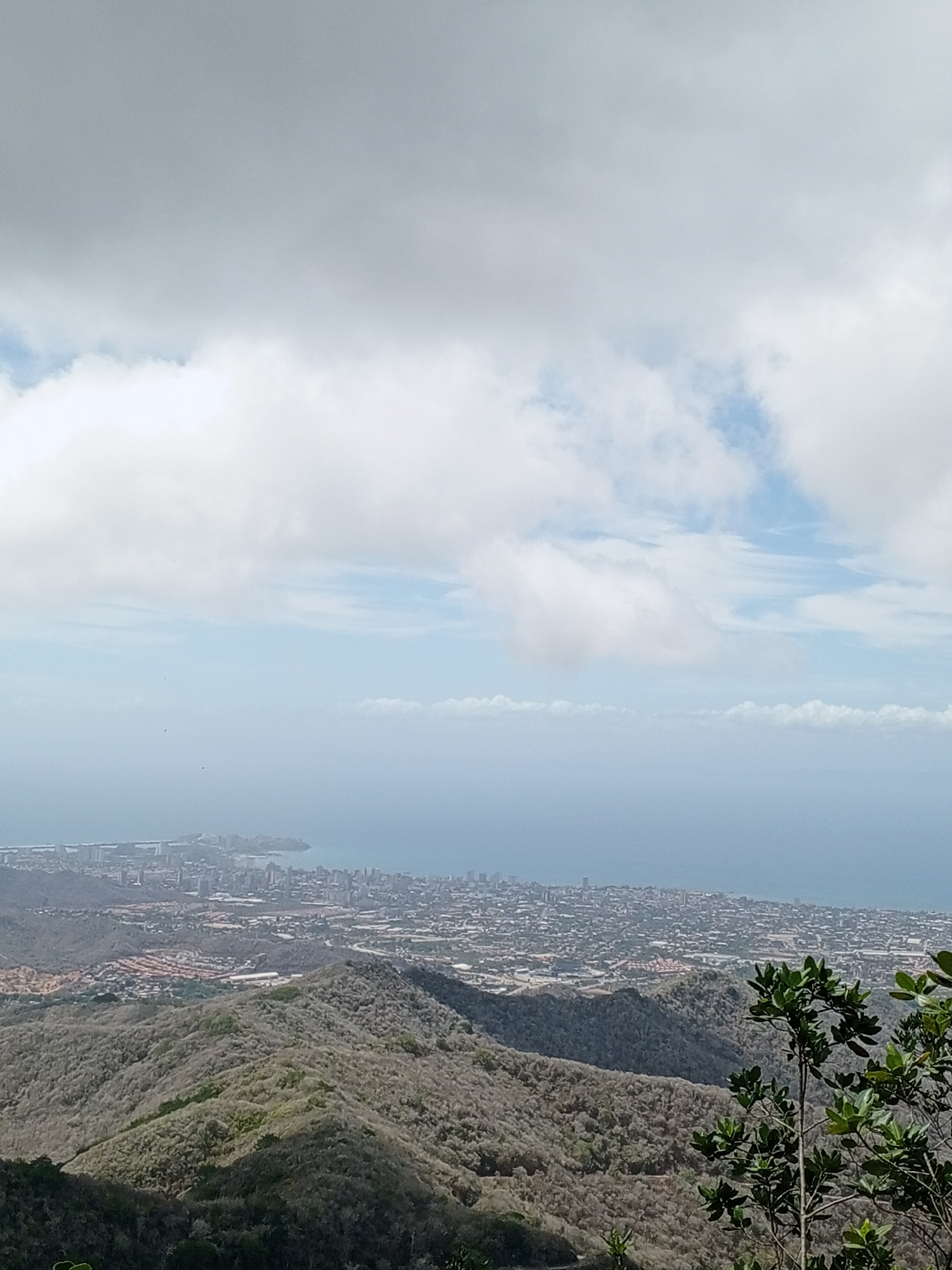 The image presents a scenic view from a mountainous region overlooking a city situated along a coastline. The cityscape, which appears to be densely populated, extends toward a peninsula that juts into the sea. The water body has a hazy appearance, likely due to atmospheric conditions.
The foreground is dominated by hills covered in sparse vegetation, with some greenery visible on the right side of the frame, suggesting closer proximity. The sky is mostly overcast, with heavy cloud cover dominating the upper portion of the image. The overall lighting suggests it is daytime.
Las Piedras, Venezuela is not visible in the image. It is not possible to confirm the location based on the visual data provided.