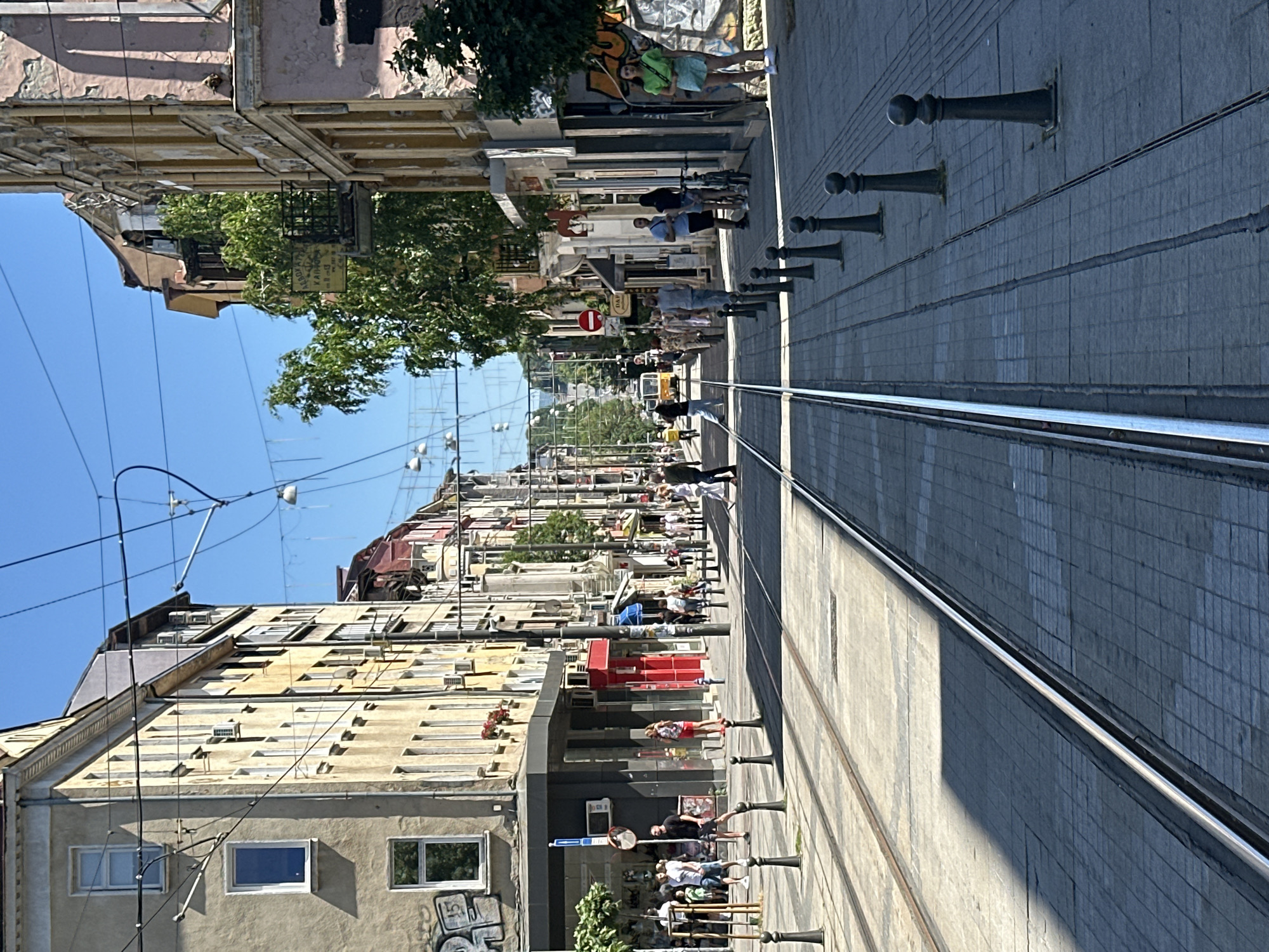 An eye-level, medium shot captures a street scene in a city with tram tracks running down the center of a paved road. On the left, a light brown building with multiple windows and some graffiti is visible, with a group of people walking on the sidewalk. On the right, a pale pink building with arched windows stands, and more people are seen on the sidewalk, some with bicycles. The buildings lining the street have multiple stories and are adorned with various architectural details. Overhead, a network of tram wires and street lights crisscrosses the sky. In the distance, a yellow tram is approaching, and the street curves and slopes upwards. The sky is a clear, bright blue.