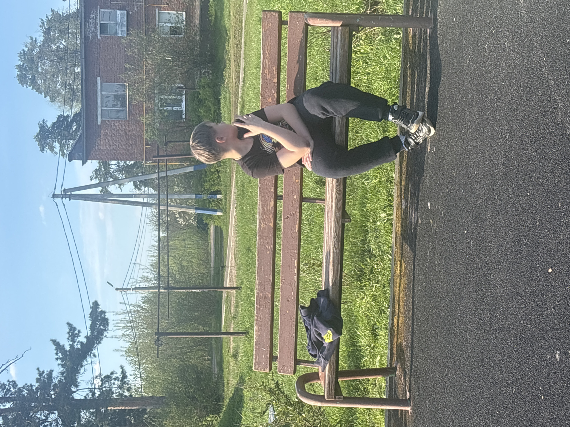 A young person is sitting cross-legged on a wooden park bench in Раздолье, Russia. The individual is wearing a dark t-shirt and dark pants with athletic shoes. Their arms are crossed in front of them. The bench is located on a grassy area adjacent to a paved surface, likely a walkway or playground area. A building is visible in the background behind some trees. Power lines and poles are also visible. A dark colored garment is also on the bench.