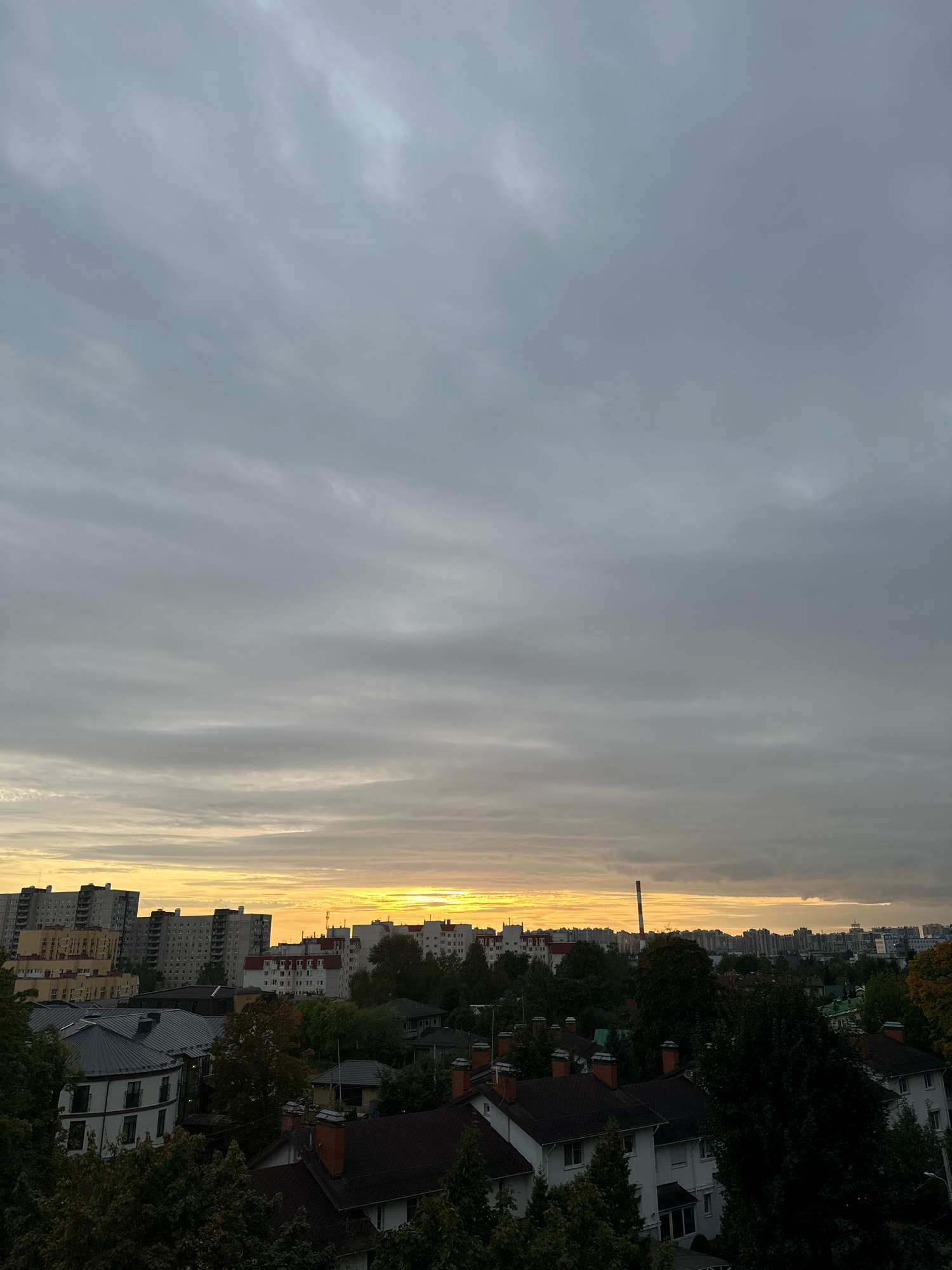 An aerial, horizontal, full shot captures a city skyline at dusk. The sky is overcast with thick, gray clouds that streak horizontally across the frame. A sliver of yellow and orange light emanates from the horizon, suggesting the sun has recently set. Below the sky, a dense urban landscape unfolds. In the foreground, the dark roofs of residential buildings with visible chimneys are prominent. Behind these, a line of lush green trees obscures much of the midground. Further back, taller apartment buildings of varying architectural styles and colors form a prominent backdrop. A red and white striped industrial chimney stands tall amongst the buildings on the right side of the frame. The overall scene depicts a transitional moment between day and night in a densely populated urban environment.