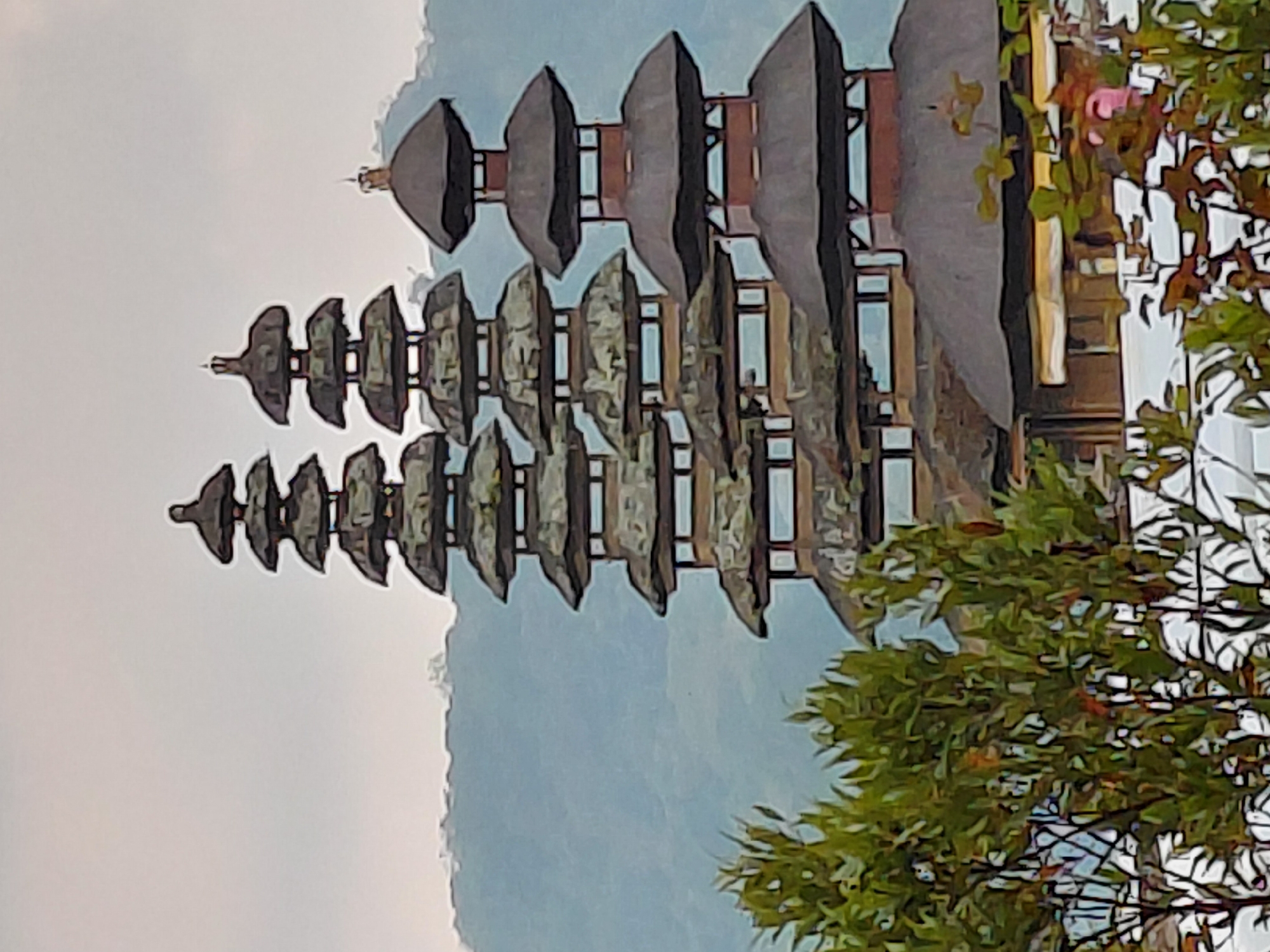 The image shows a traditional multi-tiered temple building with dark, thatched roofs against a misty, blue mountainous backdrop. The building's architecture features several stacked roofs, each with a curved, pagoda-like silhouette. The foreground is partially obscured by lush green foliage, with branches and leaves in sharp focus. The sky is overcast and pale gray. The scene appears to be a cultural or religious site, possibly a temple or shrine, given the architectural style. The location is identified as Banjar, Indonesia, which is known for its Hindu temples and serene natural surroundings.