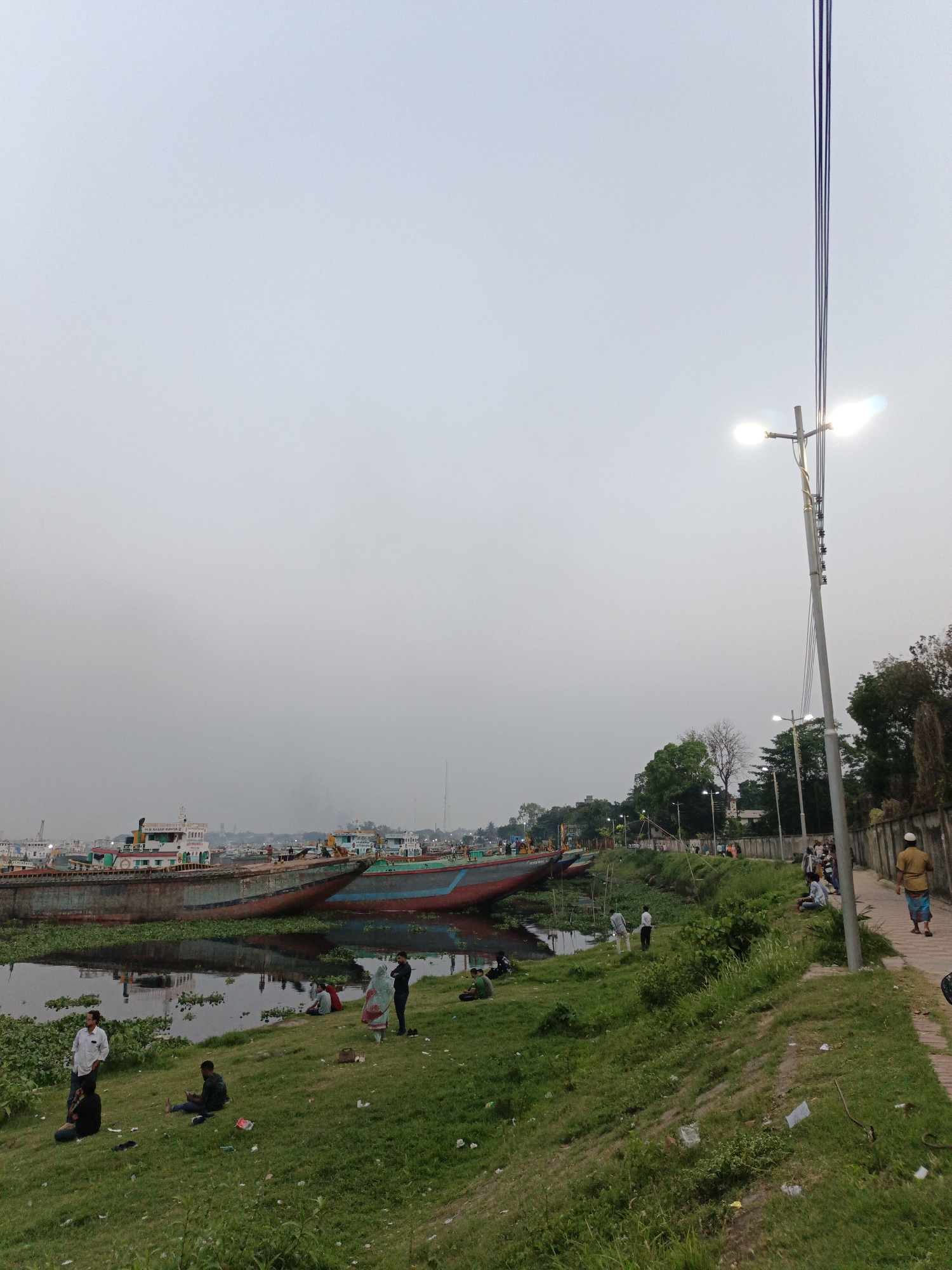 The image depicts a riverside scene in Dhaka, Bangladesh, under an overcast sky. Several large, possibly cargo, ships are docked along the muddy riverbank. A group of people are relaxing and socializing on the grassy bank in the foreground. A path runs parallel to the river, where individuals are walking. A modern street light illuminates the path. The environment suggests an urban area, with buildings and structures visible in the distance.