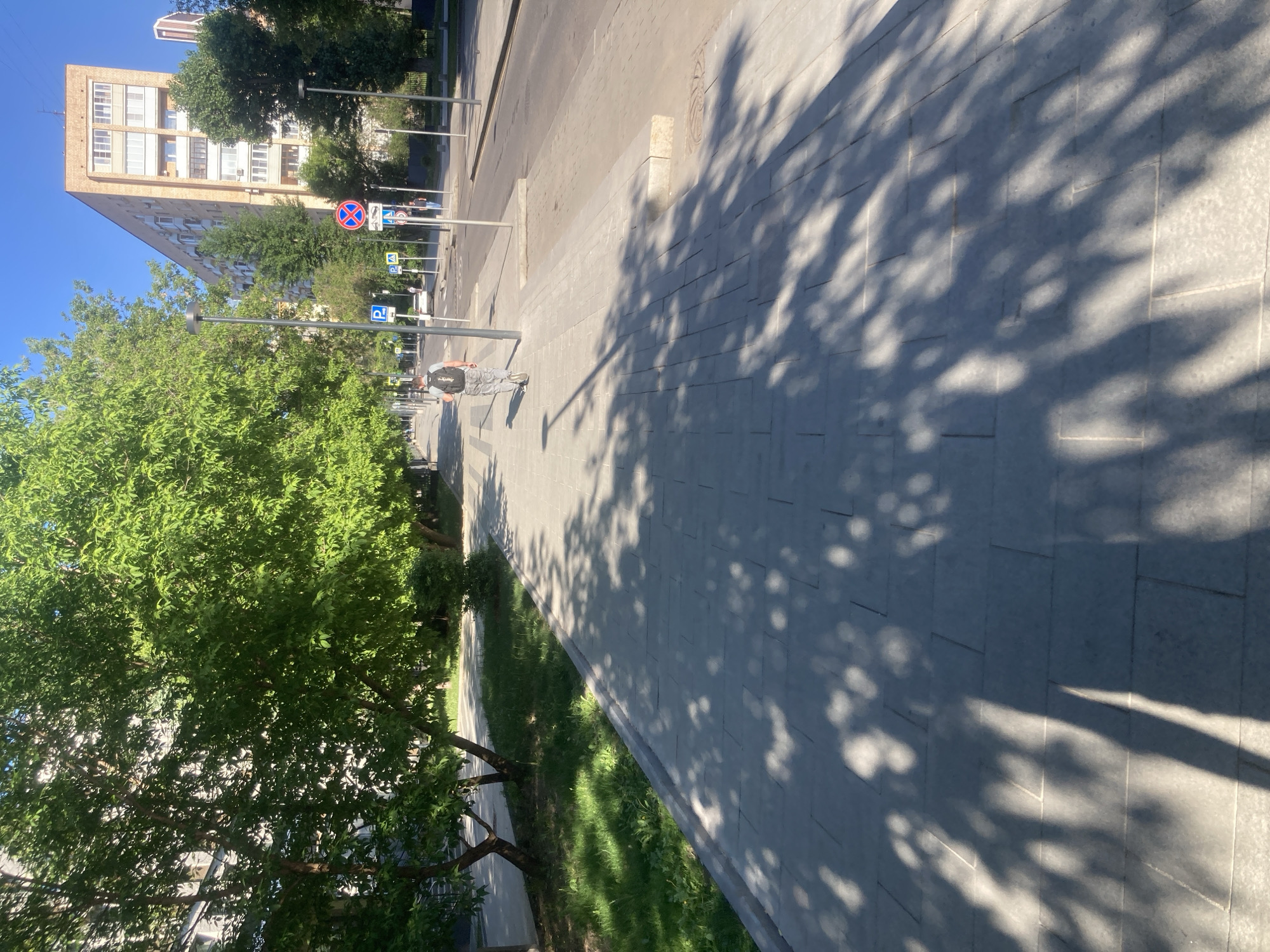 The image depicts a street scene in Moscow, Russia. A pedestrian walks along a wide sidewalk paved with grey rectangular stones. Sunlight filters through tree leaves, casting shadows on the pavement. Adjacent to the sidewalk is a strip of green grass. A road runs parallel to the sidewalk, with street lamps and parking signs visible. A tall brick building with multiple windows stands on the opposite side of the road. The sky is clear and blue.
