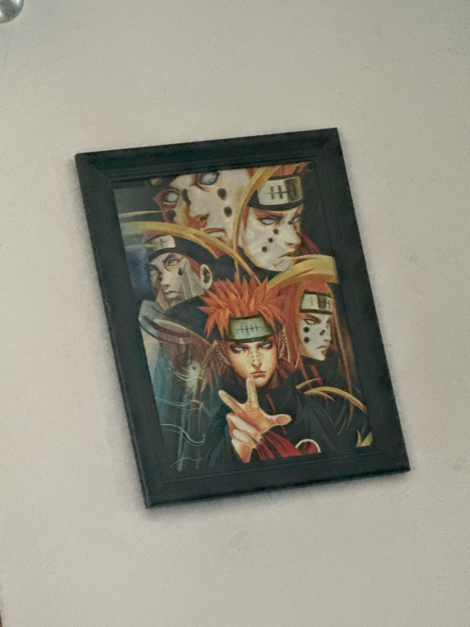 A framed artwork of six characters from the anime series Naruto, featuring Pain's Six Paths, hangs on a light-colored, textured wall. The artwork is predominantly dark with vibrant colors from the characters' hair and clothing. The characters are depicted in a close-up composition. The frame is black and simple. The image is taken at an angle, suggesting the artwork is mounted on a wall. No specific location in Mora, United States of America, can be determined from the image.
