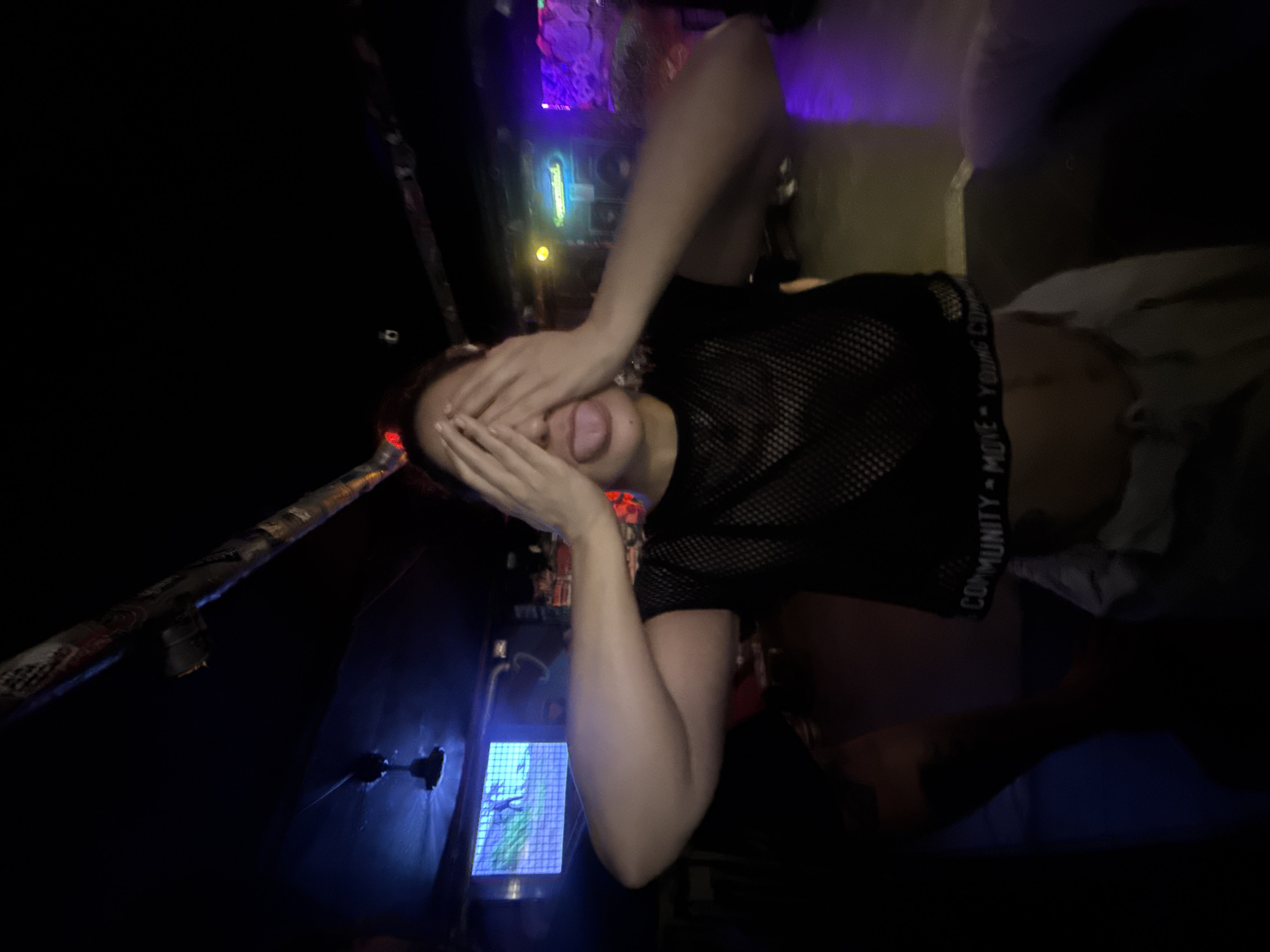 A person is lying down in a dimly lit environment, obscuring their face with their hands and sticking out their tongue. They are wearing a black mesh top with white text on the band. The background includes electronic equipment with blue and purple lighting, suggesting a nightclub or party setting. There's also a bed or seating area visible. The location is unidentifiable as Barcelona, Spain based on the image.