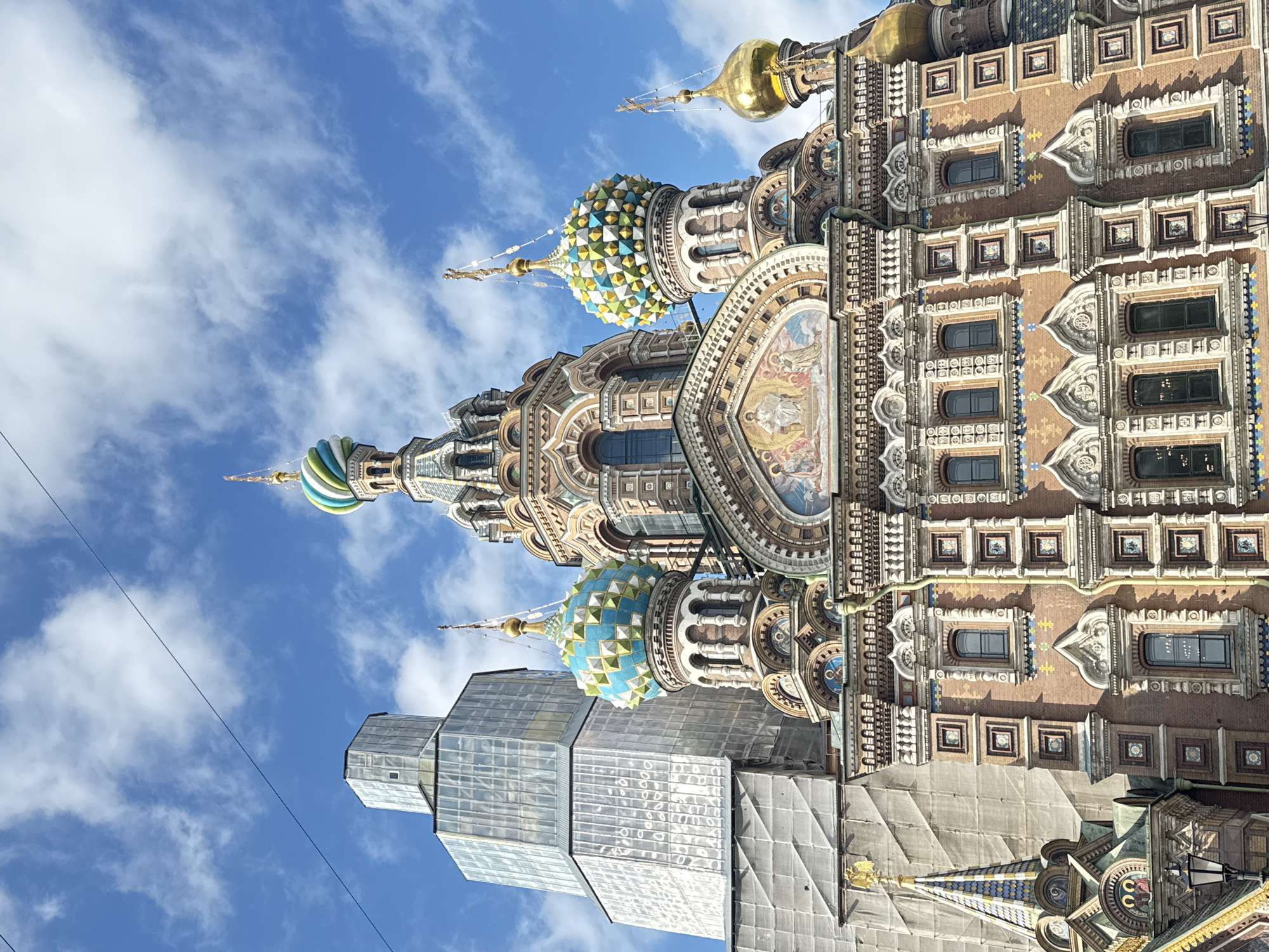 The image shows the Church of the Savior on Spilled Blood in Saint Petersburg, Russia. The church is characterized by its ornate facade, multiple domes, and colorful mosaic decorations. Part of the church is under scaffolding. The sky is visible in the background with scattered clouds.
