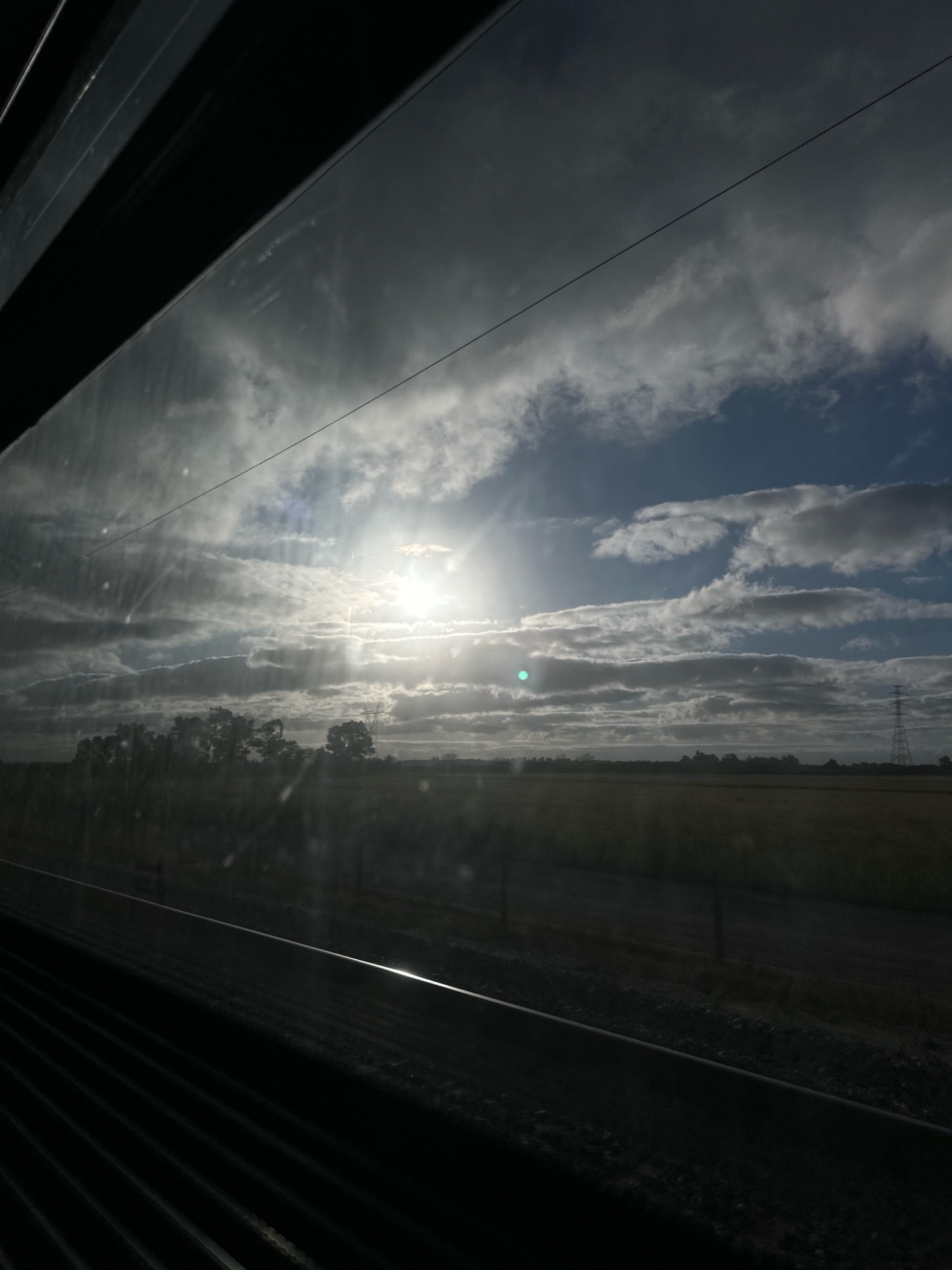 The image is a view from inside a train window, looking out at a cloudy sky with the sun shining through. The foreground is dominated by the dark interior of the train, with visible ribbed patterns and reflections on the window. The middle ground shows a silhouetted landscape with trees and a power line tower in the distance. The sky is a mix of bright white clouds and darker gray clouds, with sunbeams piercing through. The sun itself is a bright, hazy orb. The scene appears to be a rural or semi-rural area, possibly farmland, based on the flat expanse and the presence of crops. There is no indication of the specific location in Vila Nova da Rainha, Portugal, in the image itself. The image is static and depicts a landscape view.