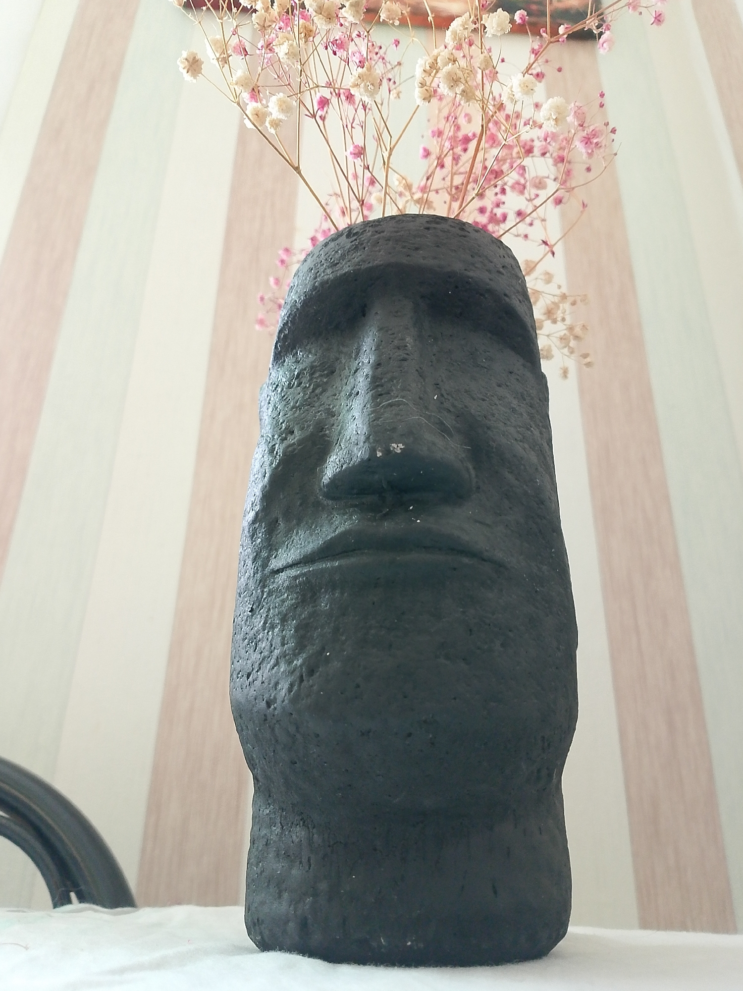 A close-up shot depicts a dark gray, textured Moai head sculpture, likely made of stone or a similar material. The sculpture has a distinct, weathered appearance. Emerging from the top of the Moai head are delicate, branched stems with small white and pink flowers, arranged as if in a vase. The sculpture rests on a white surface, possibly a tablecloth. The background consists of a wall with vertical stripes of cream and a light peach color. A portion of a black object, perhaps a chair or table leg, is visible on the left side of the frame. The location is not discernible from the image.