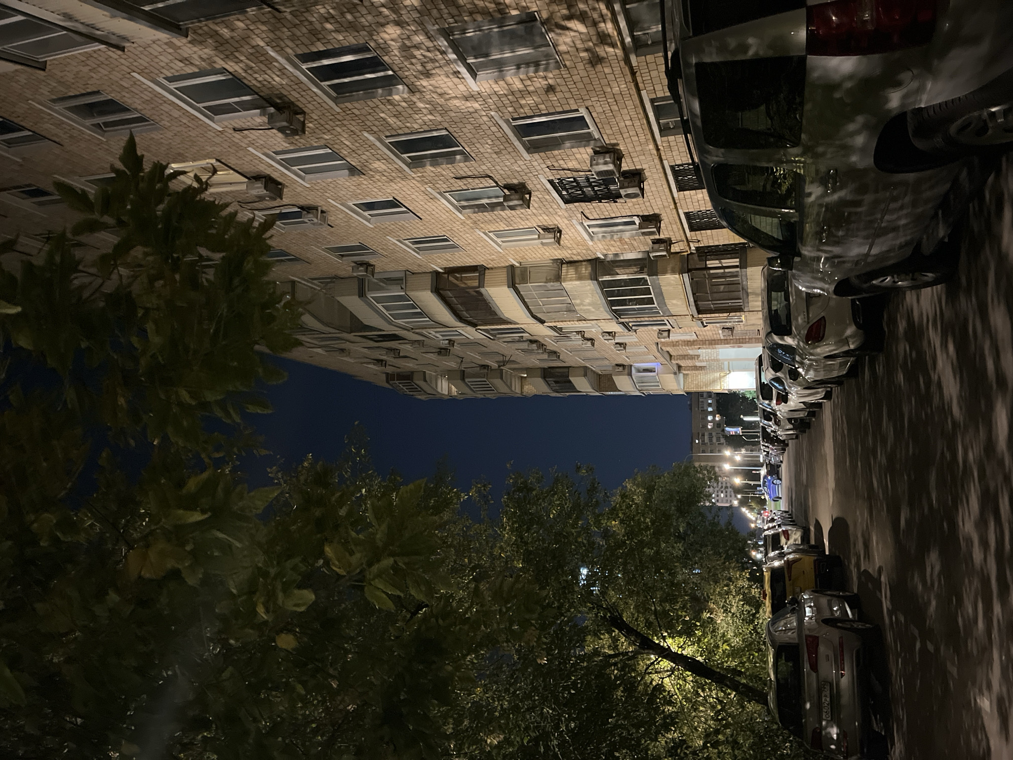 A side view of a multi-story brick apartment building is shown at night. The building has many windows, some with air conditioning units attached. Trees with green leaves hang over the street, casting shadows on the pavement. Several cars are parked along the street in front of the building, including SUVs, sedans, and a yellow hatchback. The sky is dark blue, indicating nighttime. Streetlights are visible in the distance, illuminating the street. The scene appears to be a residential area in Moscow, Russia, given the architecture of the building and the style of the cars.