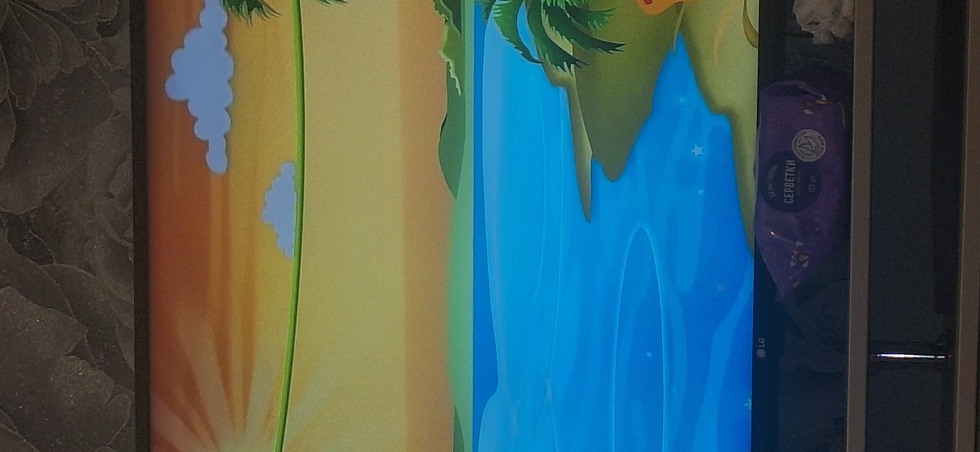 The image displays a television screen showing a cartoon landscape. The scene features a bright orange and yellow sky with stylized clouds and sun rays, suggesting a sunset or sunrise. A green vine or rope stretches horizontally across the upper portion of the frame. Below the sky is a large expanse of bright blue water, representing the sea or a large lake. Along the right edge of the water, there is a sandy or grassy shoreline with palm trees. In the foreground, resting on a shelf below the television screen, is a purple package of wet wipes with Cyrillic text that reads "СЕРБЕТКИ" (SERBETKI). The LG logo is visible on the bottom bezel of the television. The background above the television appears to be a textured wall. The image does not provide any specific information about the city of Bila Tserkva, Ukraine.