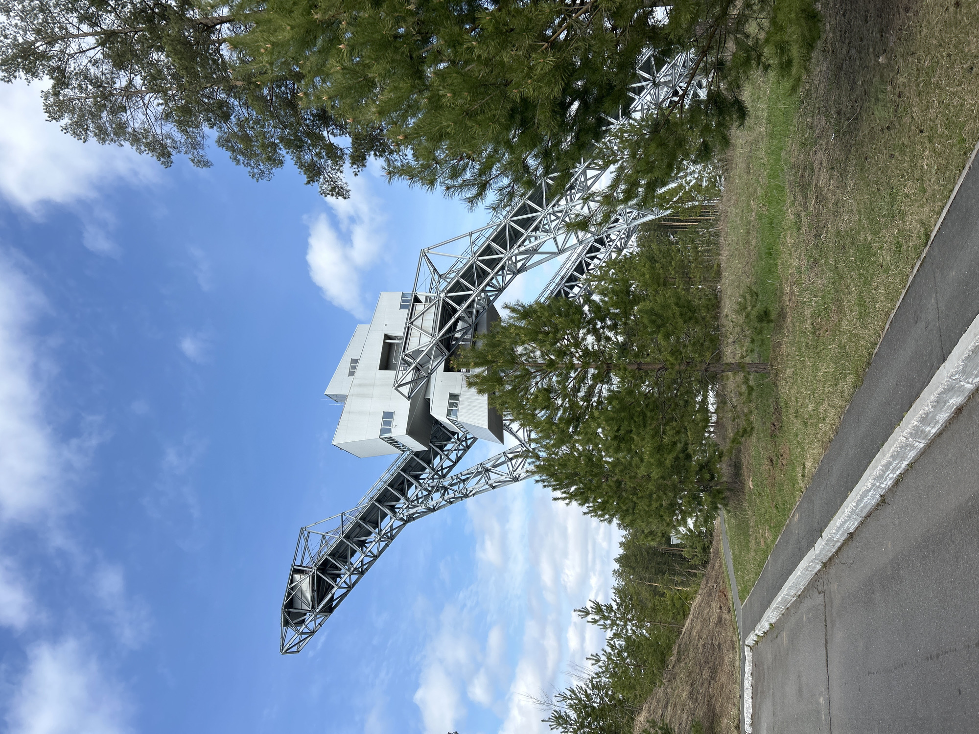 The image presents the architectural structure of the Lysvensky Ski Jumping Complex, located in the city of Прикамский, Russia. The structure consists of a white, multi-tiered building perched atop a metal framework that descends towards the ground. The framework is constructed of interwoven metal beams, forming a grid-like design. The sky is visible behind the structure, dotted with scattered clouds. Green trees frame the scene, both at the top and along the right side, creating a natural border. A paved road is visible at the bottom right, with a white barrier marking its edge. The perspective is angled upwards, showcasing the height and scale of the ski jumping complex.